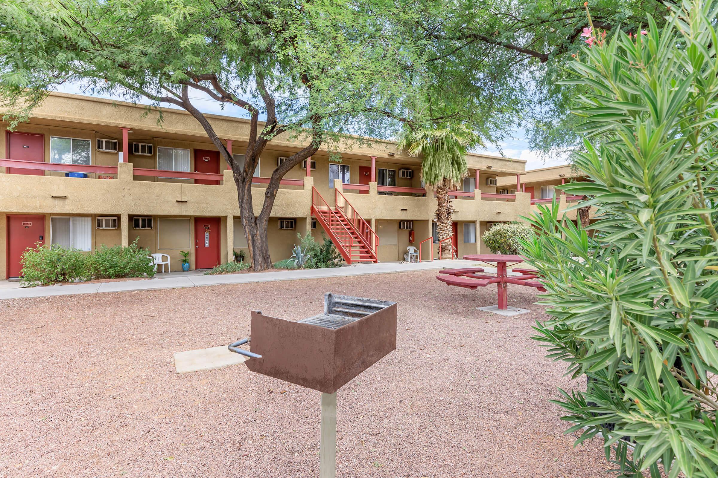 A courtyard view of a motel with two-story beige buildings, red doors, and balconies. There is a picnic area with a red table and a barbecue grill. Surrounding greenery includes shrubs and trees, providing a shaded and inviting atmosphere. The ground is covered in gravel.