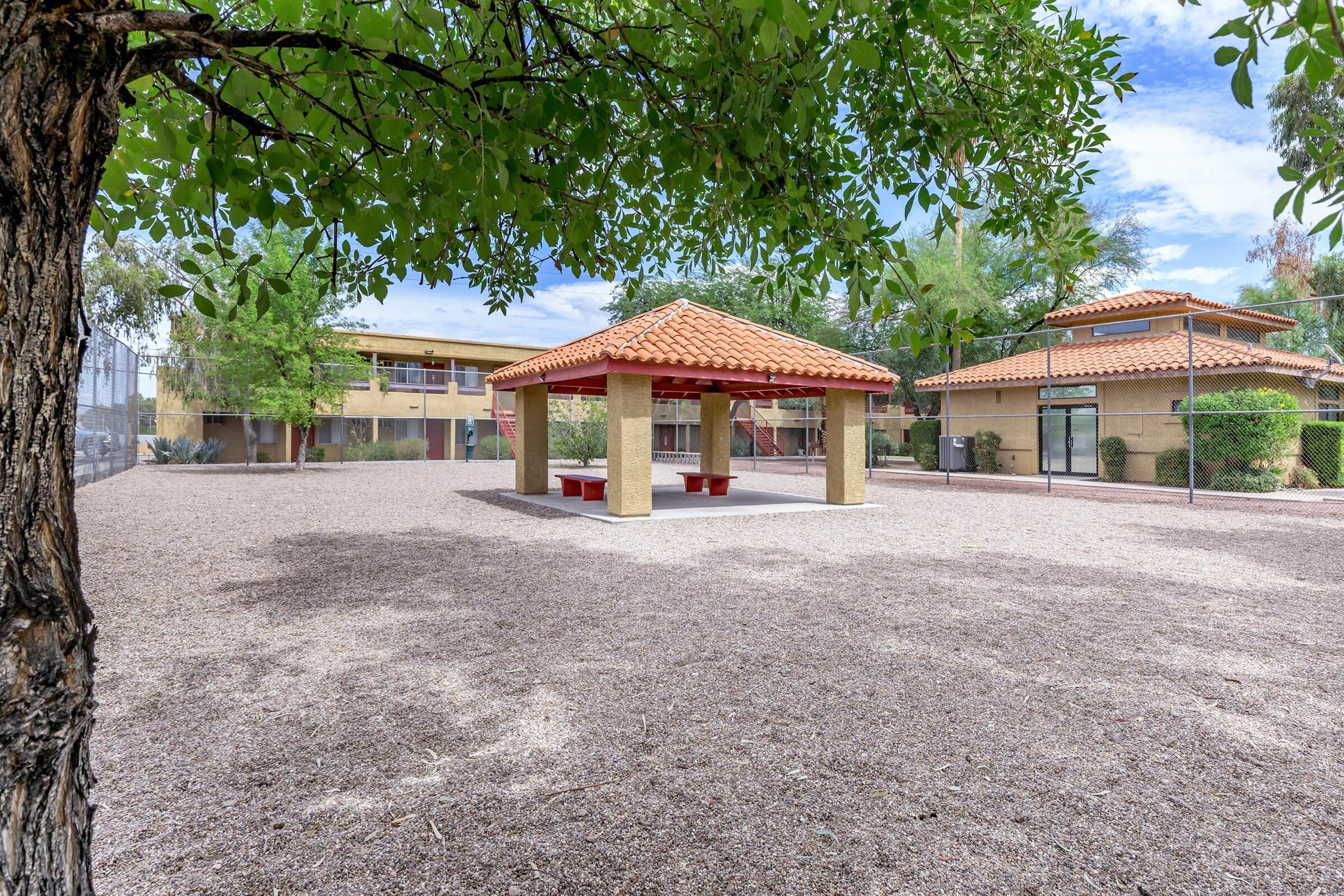 A shaded gazebo with a tiled roof sits in a gravel area surrounded by trees and buildings. The scene is bright with clear skies, showcasing a peaceful outdoor space ideal for relaxation or gatherings.