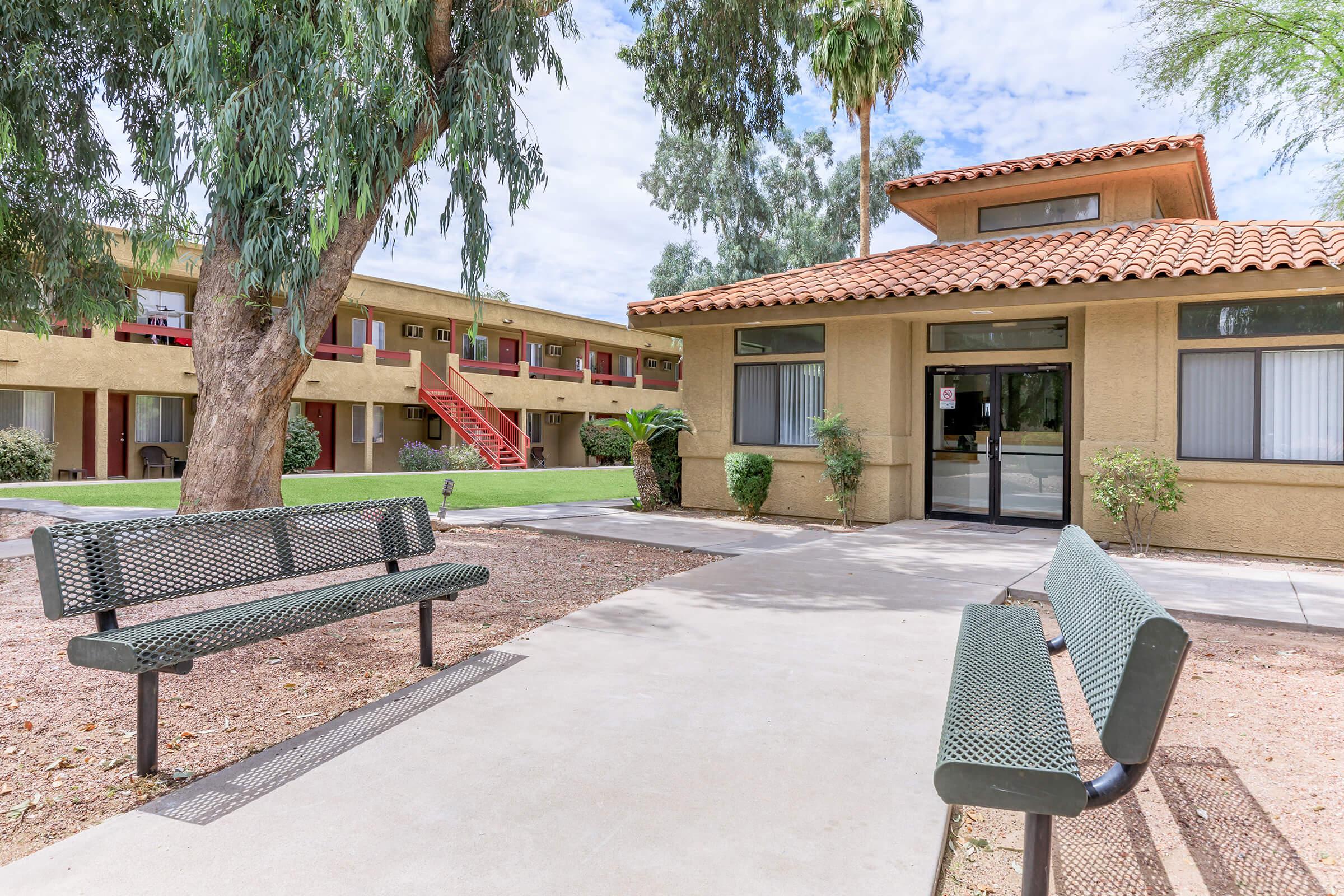 A view of the entrance to an apartment complex featuring a small office building with a tiled roof, surrounded by well-maintained landscaping. Two benches are positioned on a concrete path, and there are palm trees and shrubs in the vicinity, creating a welcoming atmosphere.