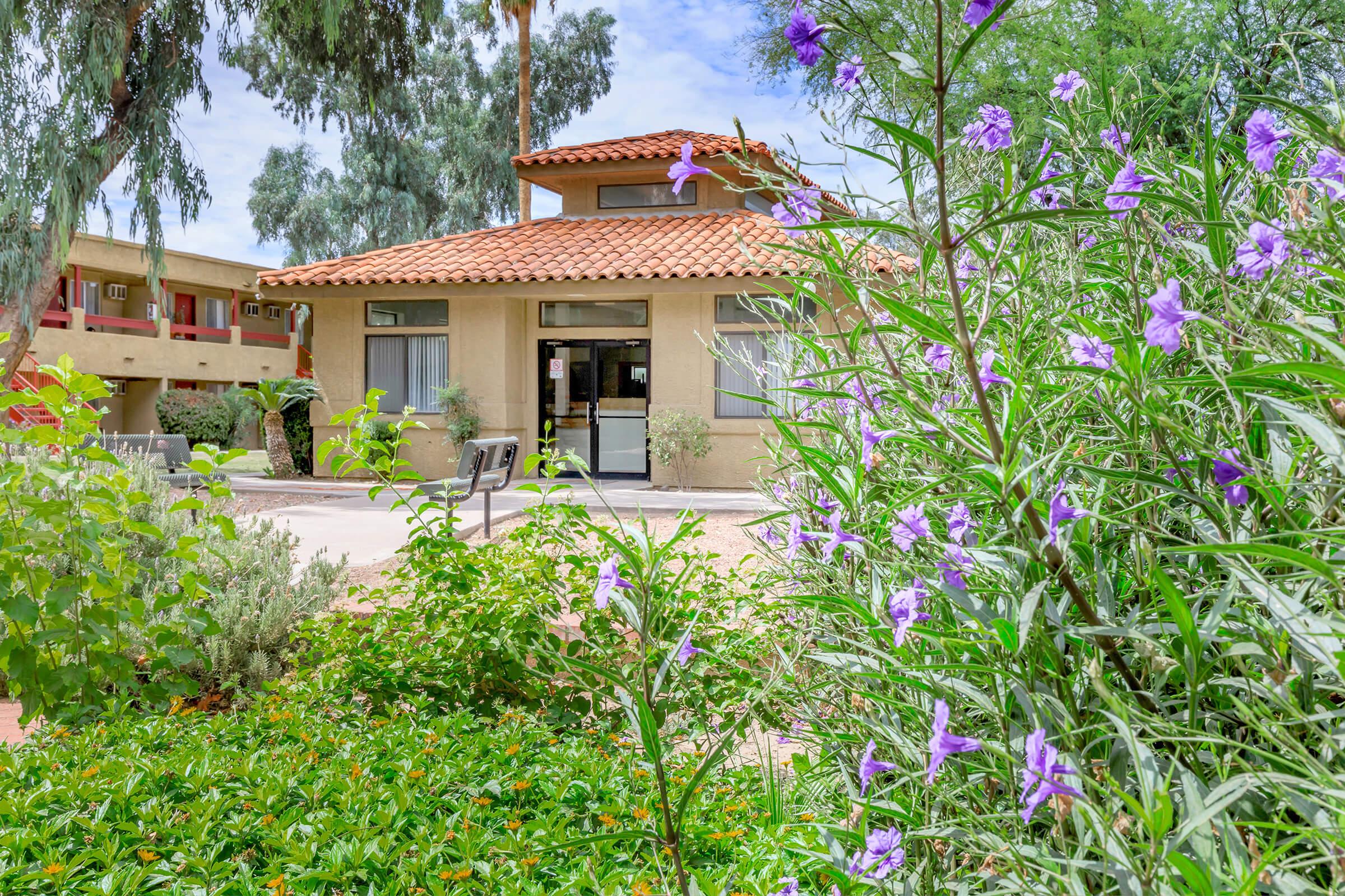 A landscaped area with flowering plants and greenery in the foreground, leading to a building with a red tile roof. The building features large windows and is surrounded by trees. In the background, additional structures are visible, creating a serene outdoor environment.