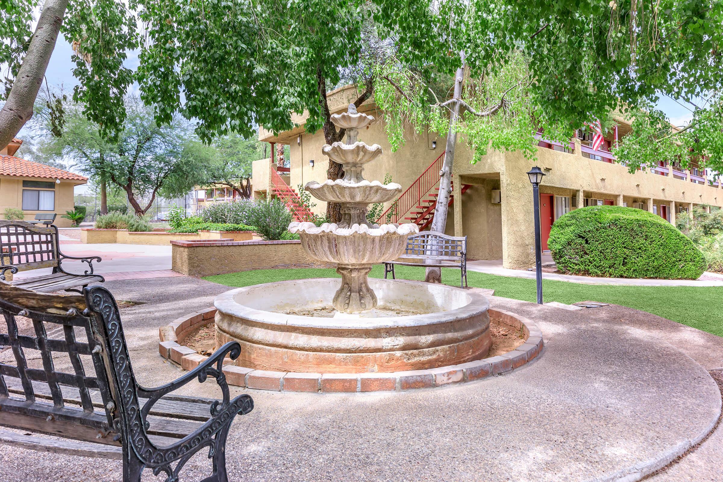 A tranquil courtyard featuring a tiered stone fountain surrounded by lush greenery. Nearby, there are benches for seating and staircases leading to upper-level buildings. The setting has a peaceful ambiance, ideal for relaxation or socializing.
