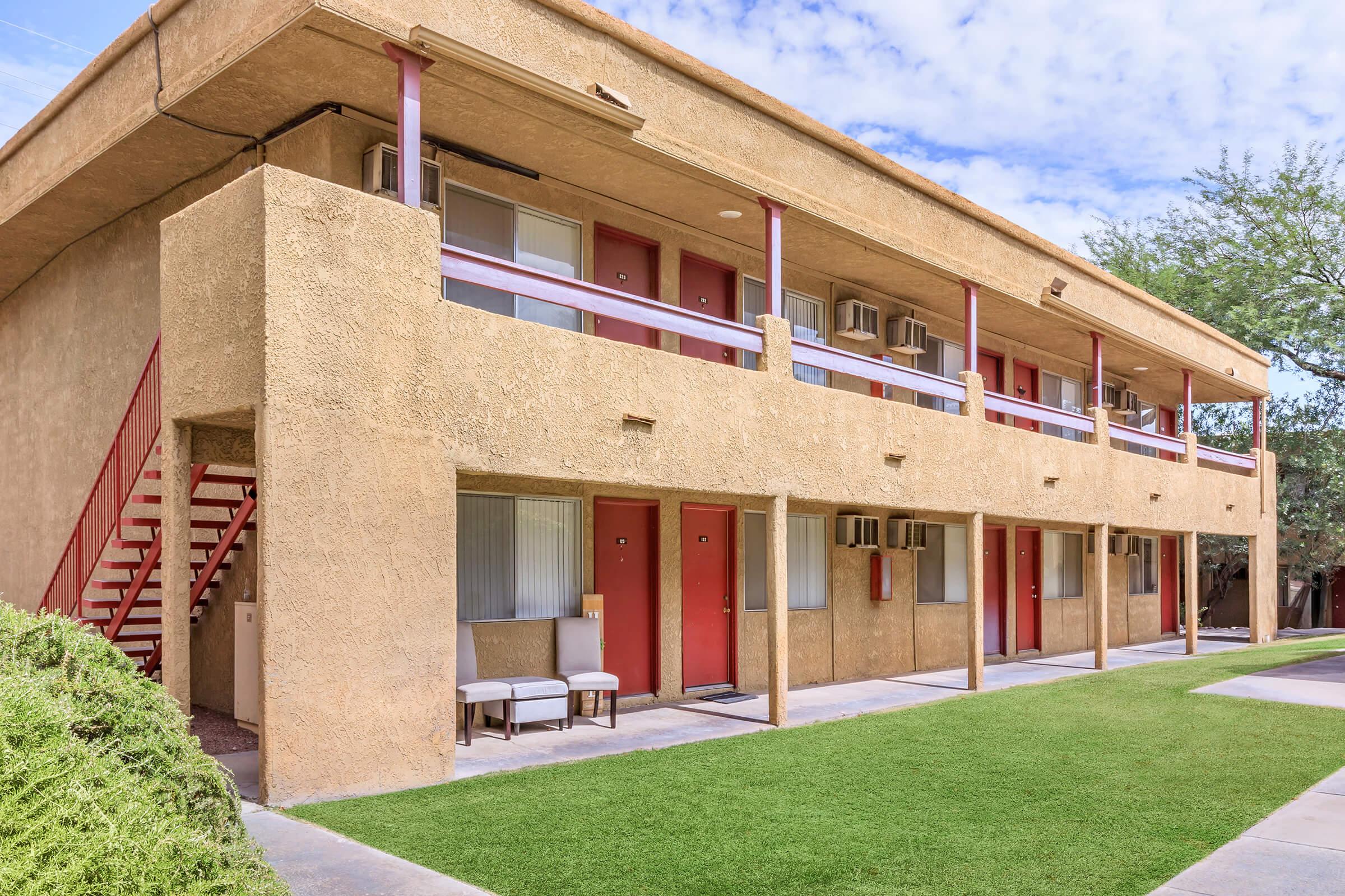 Exterior view of a two-story apartment building with a stucco facade. The building features red doors, air conditioning units, and a stairway leading to the upper level. A small seating area with two chairs is visible in front of the building, surrounded by green grass and landscaping. The sky is partly cloudy.