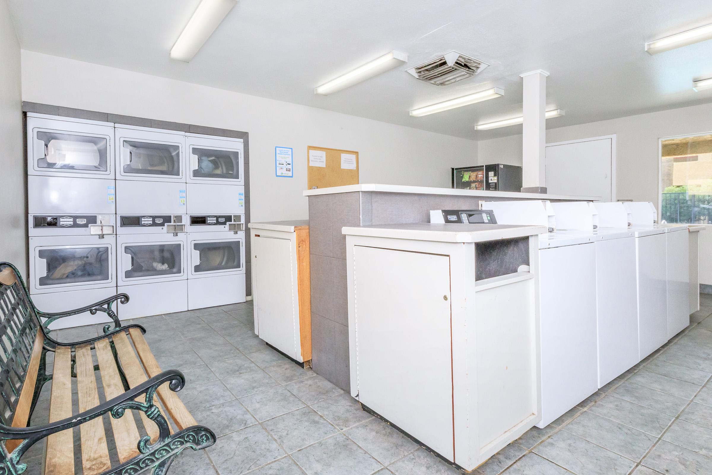 Interior of a laundry room featuring white washing machines and dryers along one wall, a counter with additional machines, and a bench for seating. Bright lighting illuminates the clean, tiled space with a neutral color scheme.