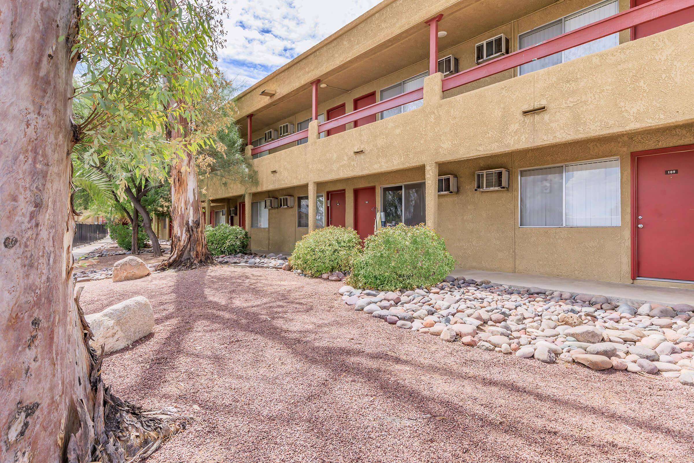 Ground-level view of a building with a tan exterior featuring red doors. Surrounding the building are landscaped areas with shrubs and large rocks, set against a backdrop of trees. The sky is partly cloudy, and there are air conditioning units under the windows.