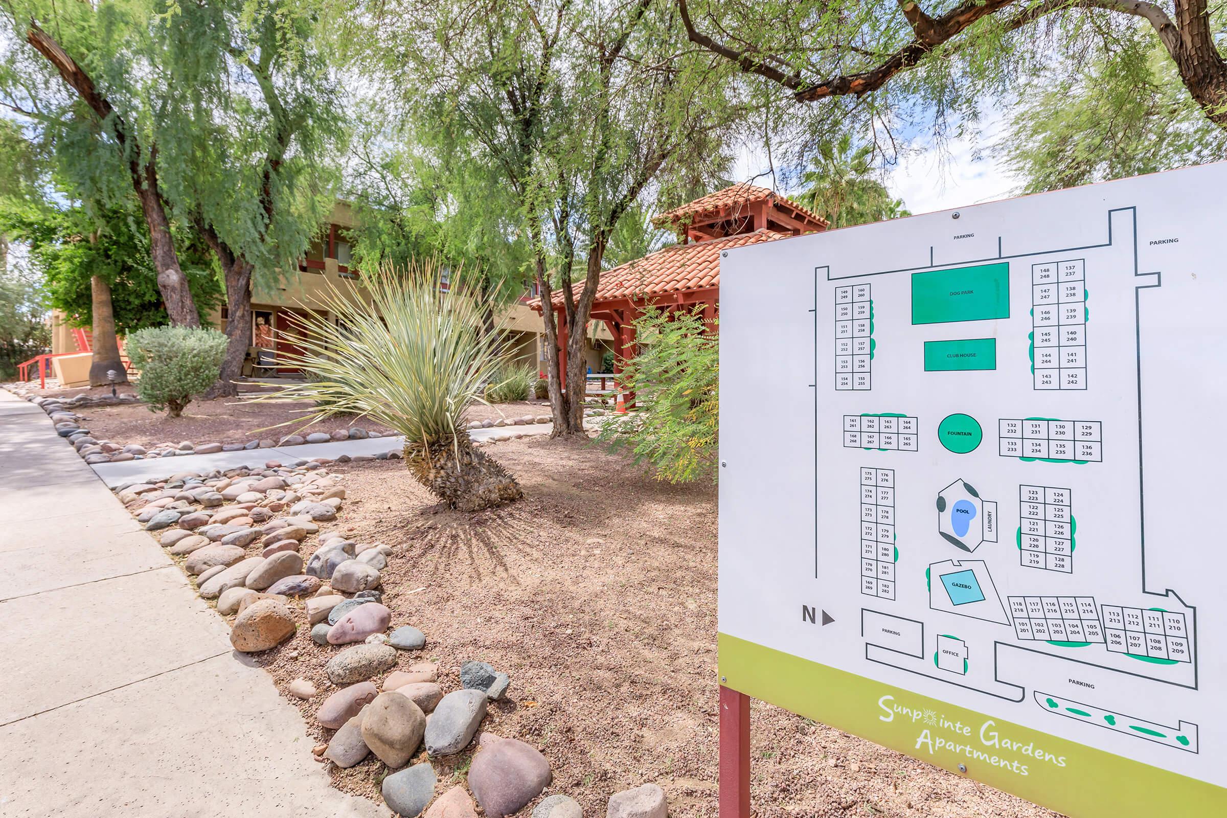 Map showing the layout of Sympatico Gardens Apartments, with nearby landscaping featuring rocks, shrubs, and trees. The two-story building can be seen behind the sign, which also includes a key to different apartment locations and amenities within the complex.