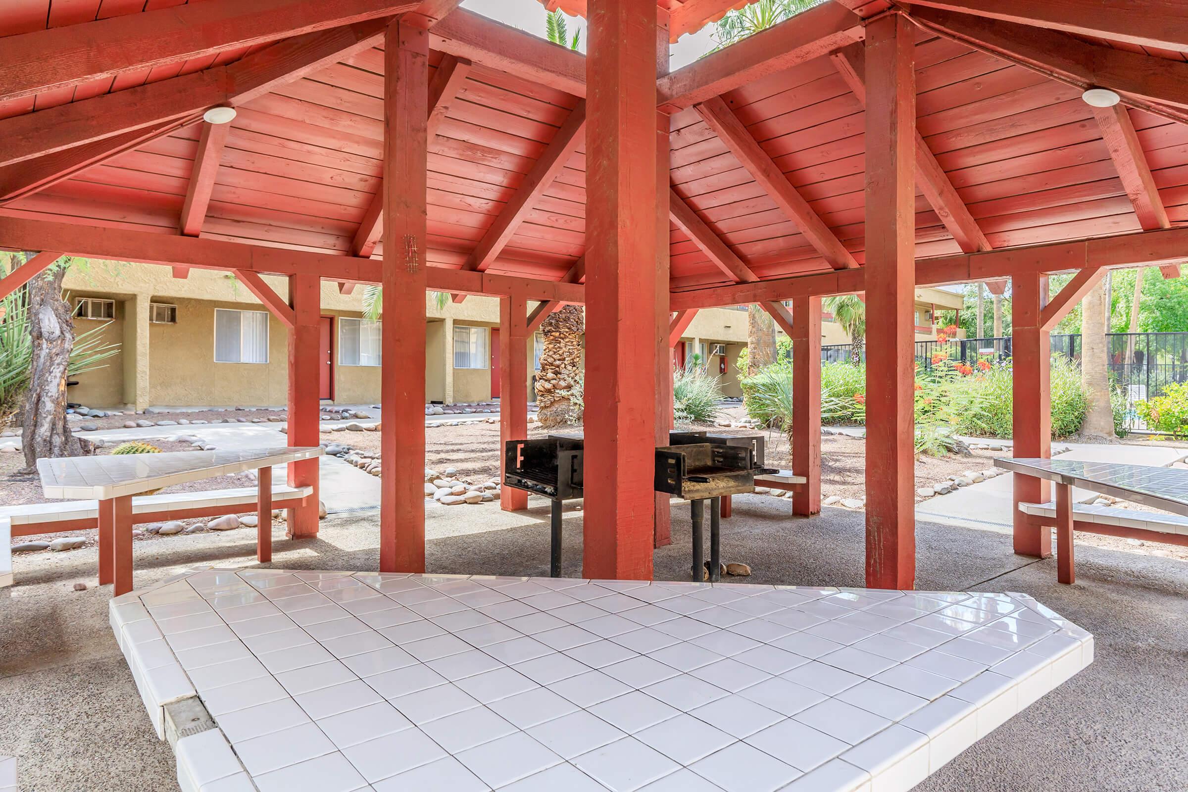 An outdoor pavilion with a red wooden structure featuring a central grilling area and several white tile-topped tables. Surrounding the pavilion are small palm trees and landscaped areas. The background shows a residential building with beige walls. 