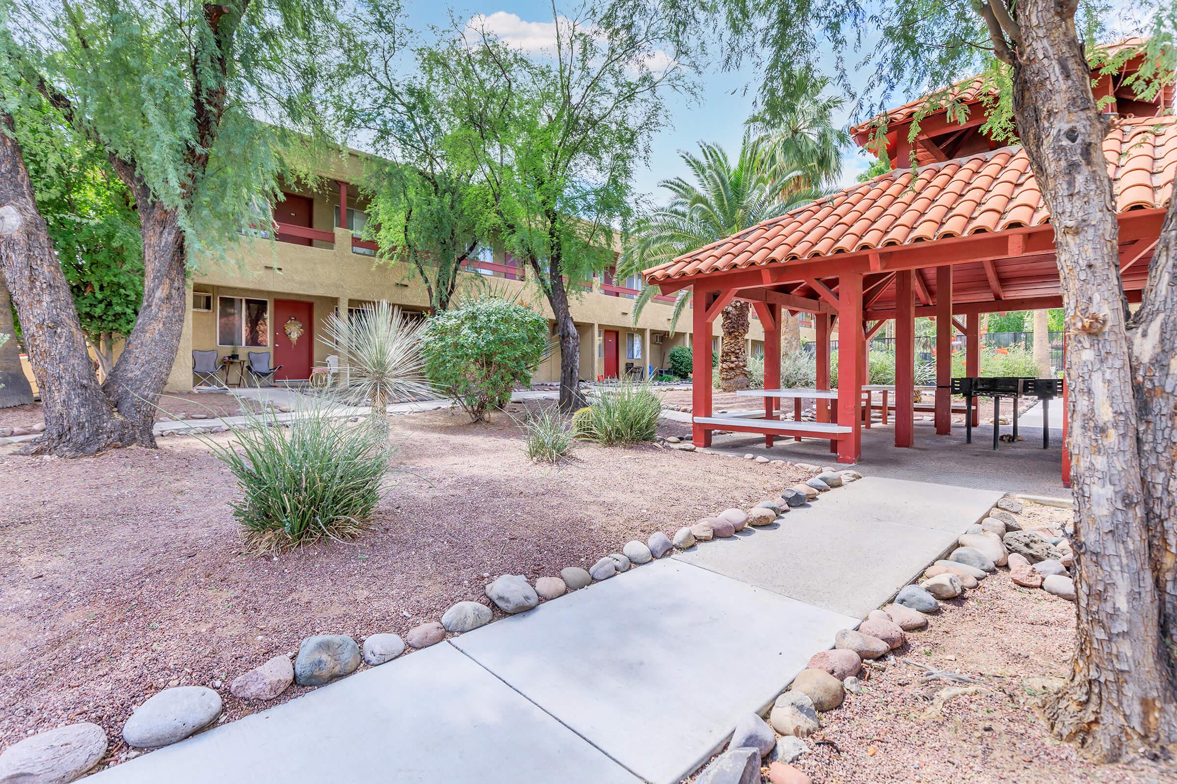 A scenic view of a landscaped courtyard featuring a stone pathway leading to a red-roofed gazebo. Surrounding the gazebo are shrubs, plants, and palm trees. In the background, two-story buildings are visible, creating a serene, inviting outdoor space.