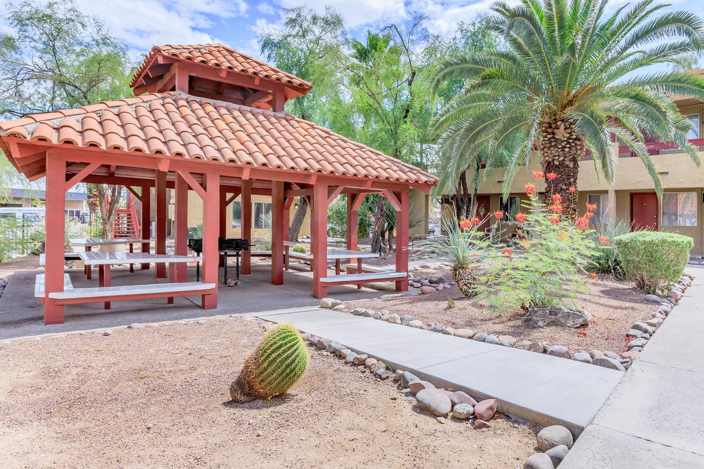 A shaded gazebo with a red tiled roof, surrounded by landscaped shrubs, palm trees, and gravel pathways. The area features picnic tables beneath the gazebo, offering a place for outdoor gatherings. A small cactus is visible in the foreground, adding to the desert ambiance.
