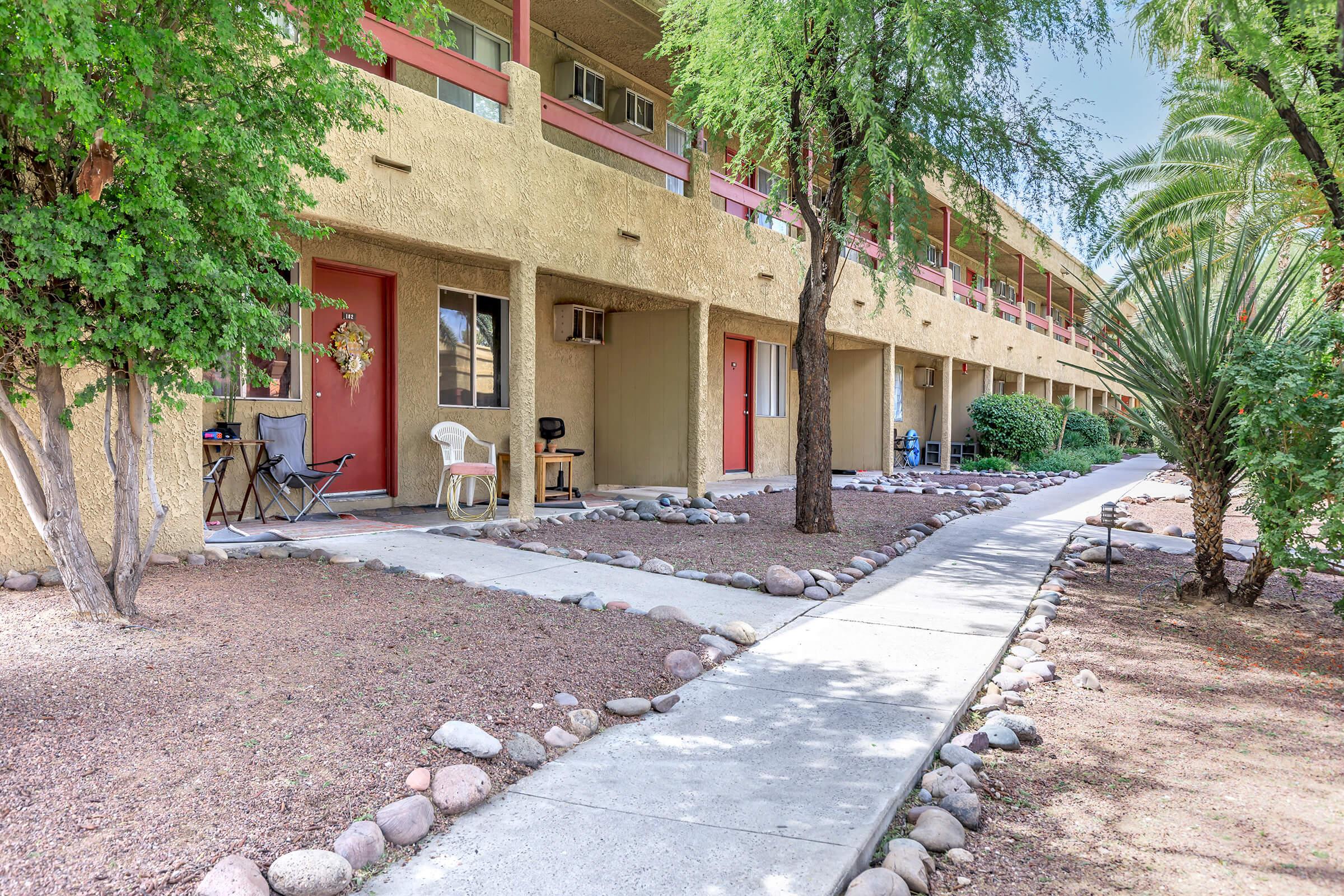 View of a walkway lined with rocks leading to a series of residential units in an apartment complex. Each unit features a front door, with a small seating area and decorative elements. Lush greenery including bushes and palm trees surrounds the area, enhancing the outdoor ambiance.