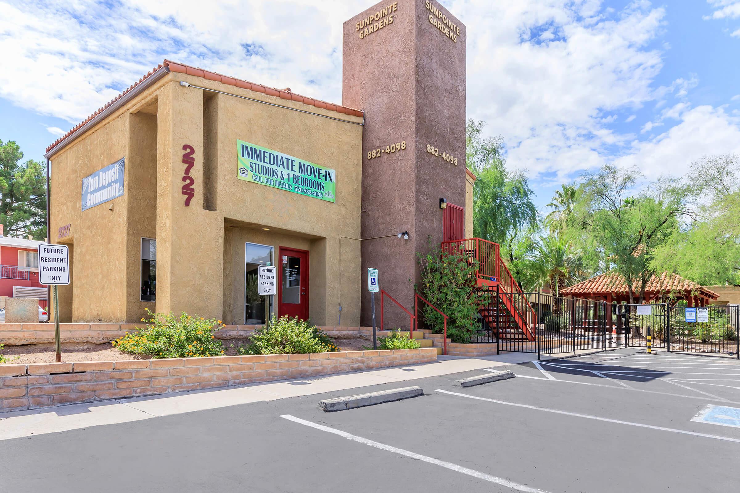 A two-story apartment building with a staircase on the side, featuring a beige exterior and a red door. A large sign on the front indicates "IMMEDIATE MOVE IN" with additional text. Surrounding the building are landscaped areas with shrubs and a parking lot visible in the foreground.