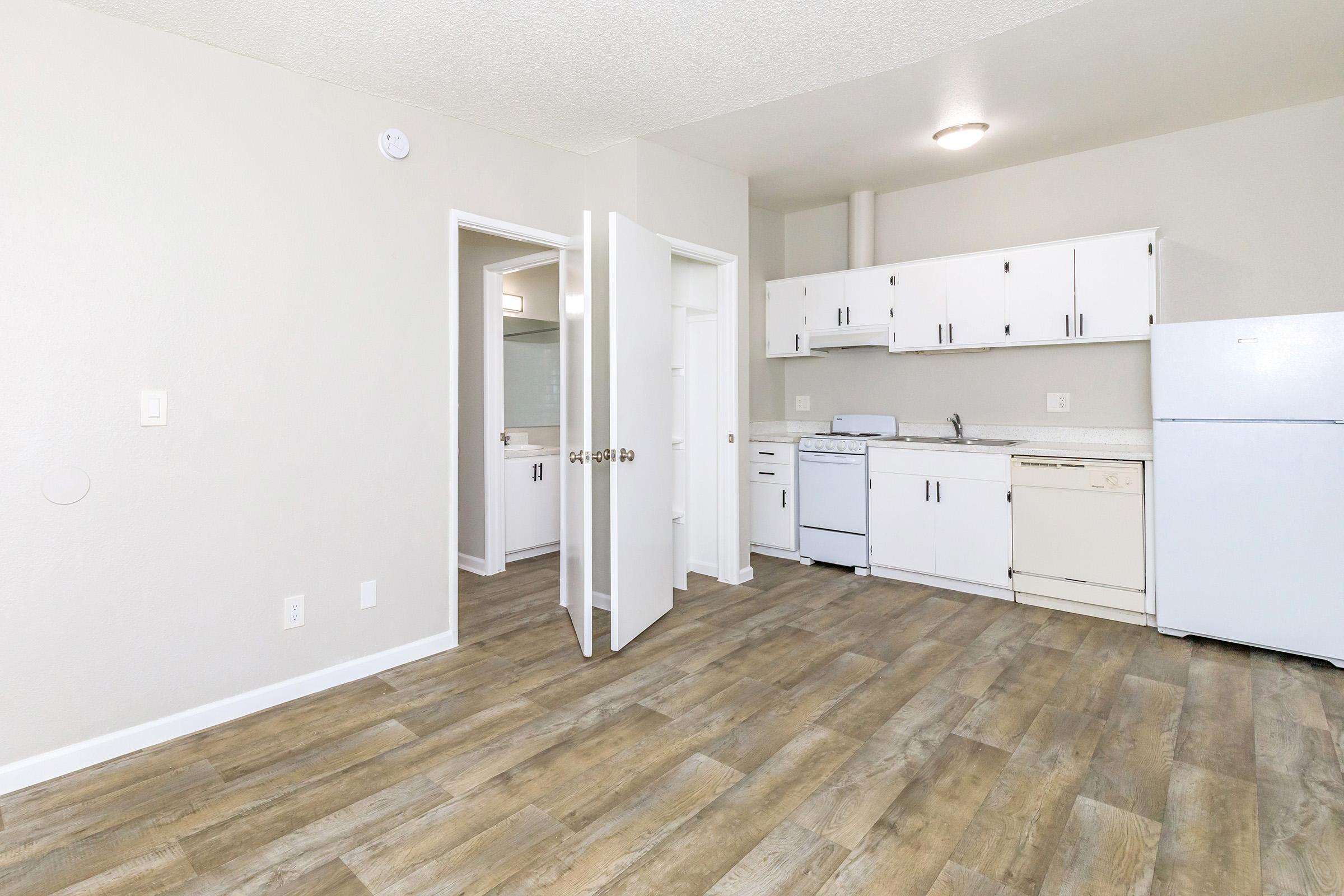 A small, modern kitchen and living area featuring wood-effect flooring, white cabinetry, a refrigerator, and a stove. Two open doors show a bathroom and a closet. The walls are light-colored, and the space is bright and airy, suggesting a recently renovated apartment.