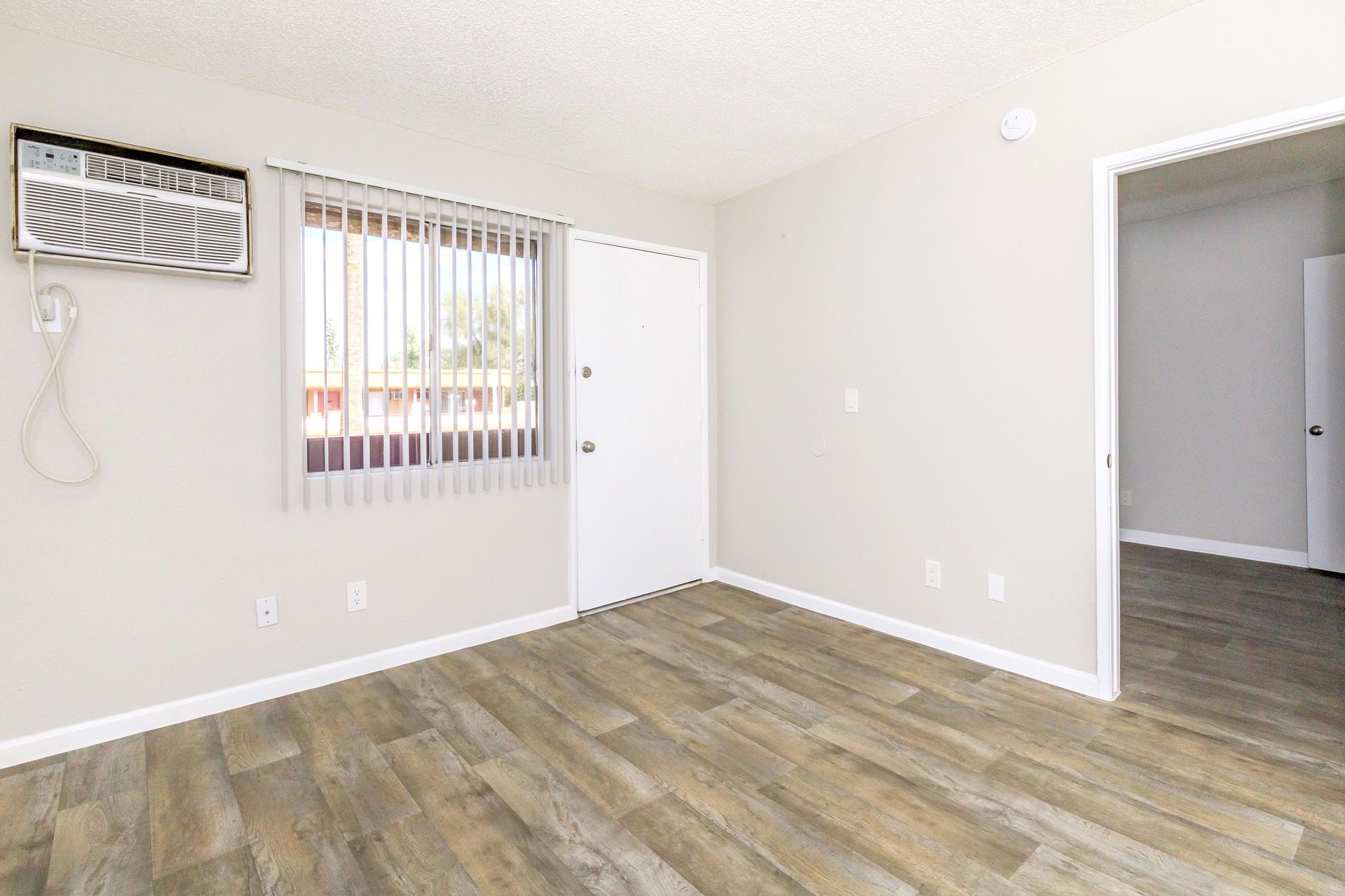A small, empty room with a light-colored wall, wooden flooring, and a wall-mounted air conditioning unit. There is a window with vertical blinds, an entrance door with a doorknob, and a glimpse of a room through an open door on the right. The space is well-lit and features a minimalist design.