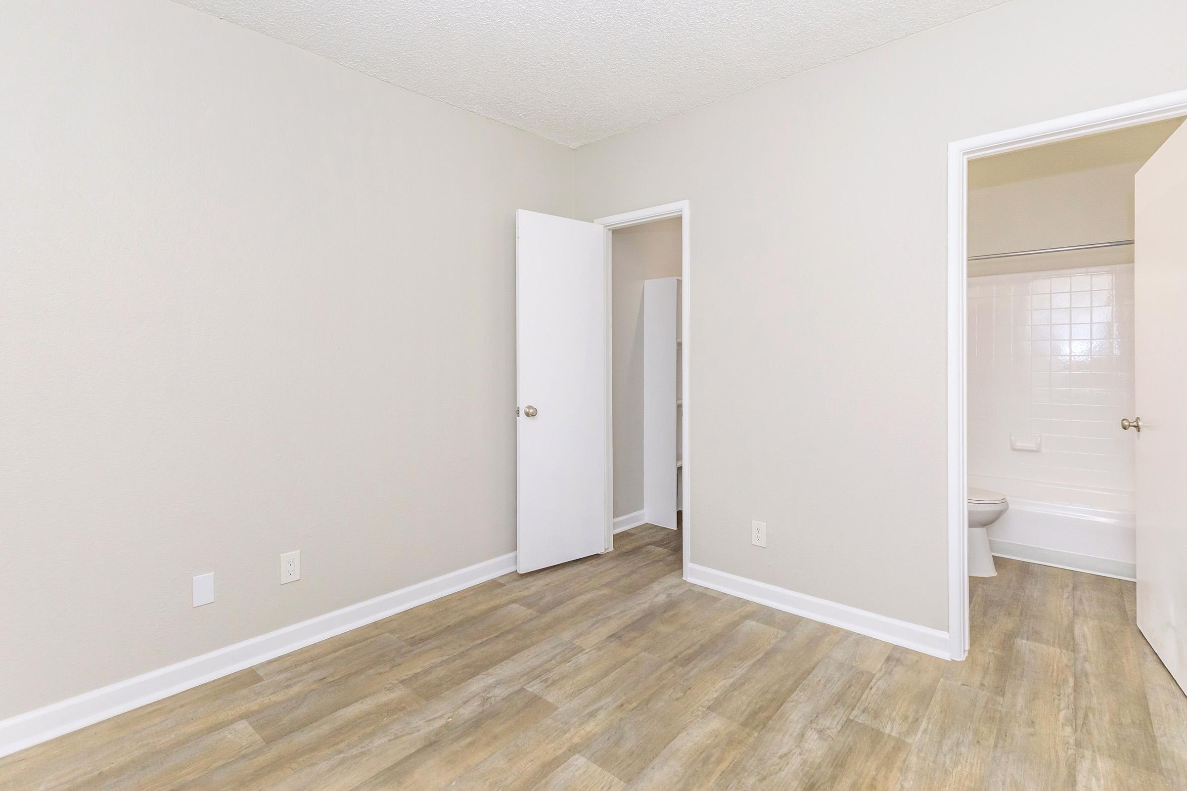 A minimalist room with light gray walls and a wooden floor. The space features a white door leading to a small closet, another white door that opens to a bathroom, and an overall clean and bright appearance, ideal for personal or rental use.