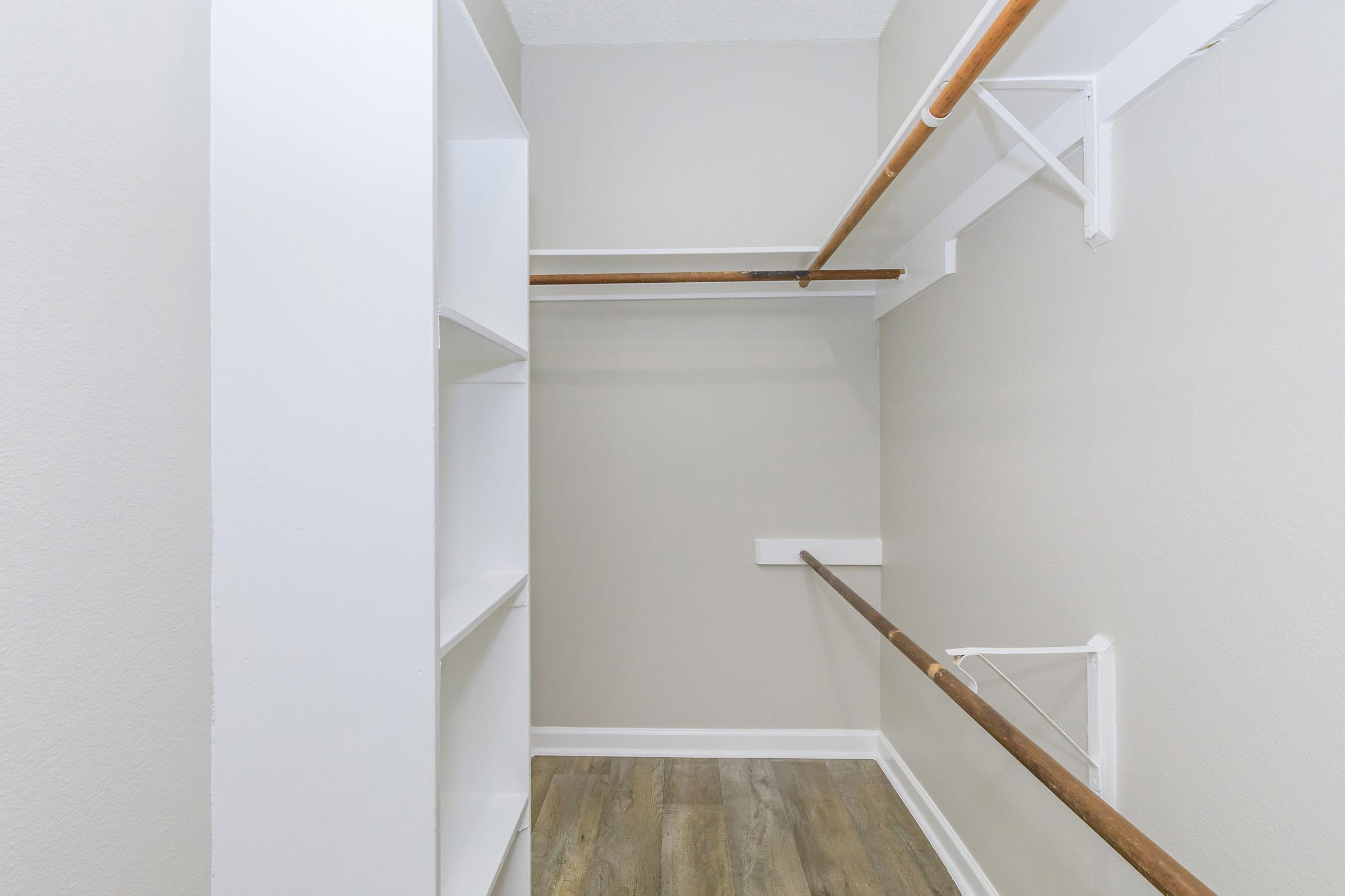 A well-organized closet with white shelves on the left and two wooden hanging rods for clothes on the right. The walls are painted in a light gray color, and the flooring is a wood laminate. The space appears clean and spacious, suitable for storage and organization.