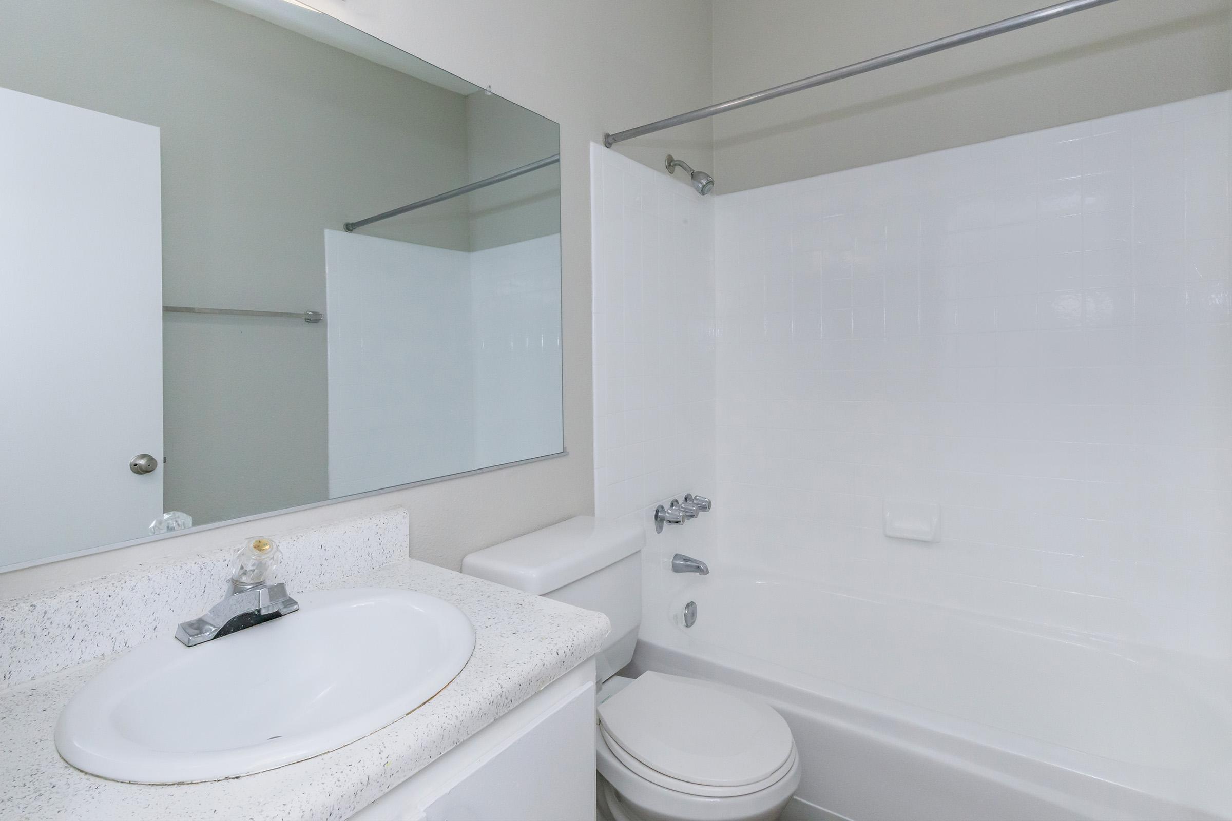 A clean, modern bathroom featuring a white bathtub, a toilet, a sink with a simple countertop, and a large mirror. The walls are painted in a neutral tone, and there is a shower curtain on the side. The overall ambiance is bright and uncluttered.