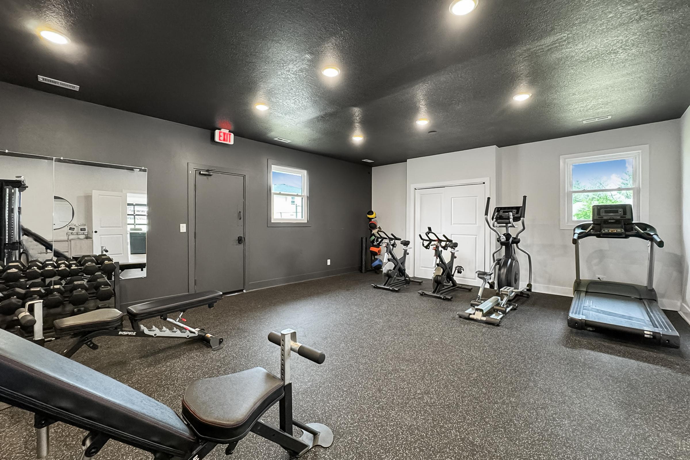 Interior view of a modern gym featuring exercise equipment. There are free weights on a bench, an elliptical machine, a treadmill, and exercise bikes, all against a dark wall with bright overhead lighting. Windows provide natural light, enhancing the workout space.