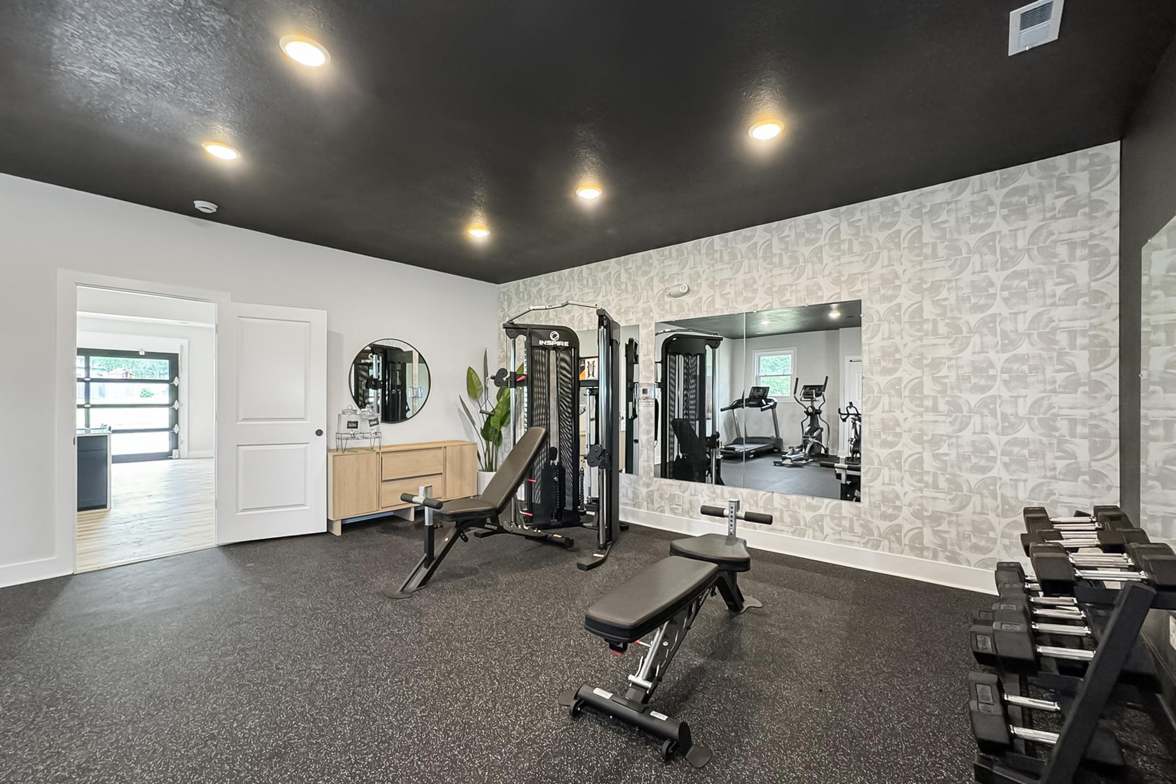 A modern home gym featuring a weight bench, adjustable weight machine, and a mirror on the wall. The floor is covered with black rubber matting, and there is a set of dumbbells in the corner. Natural light streams in through an open door, enhancing the sleek, contemporary design.