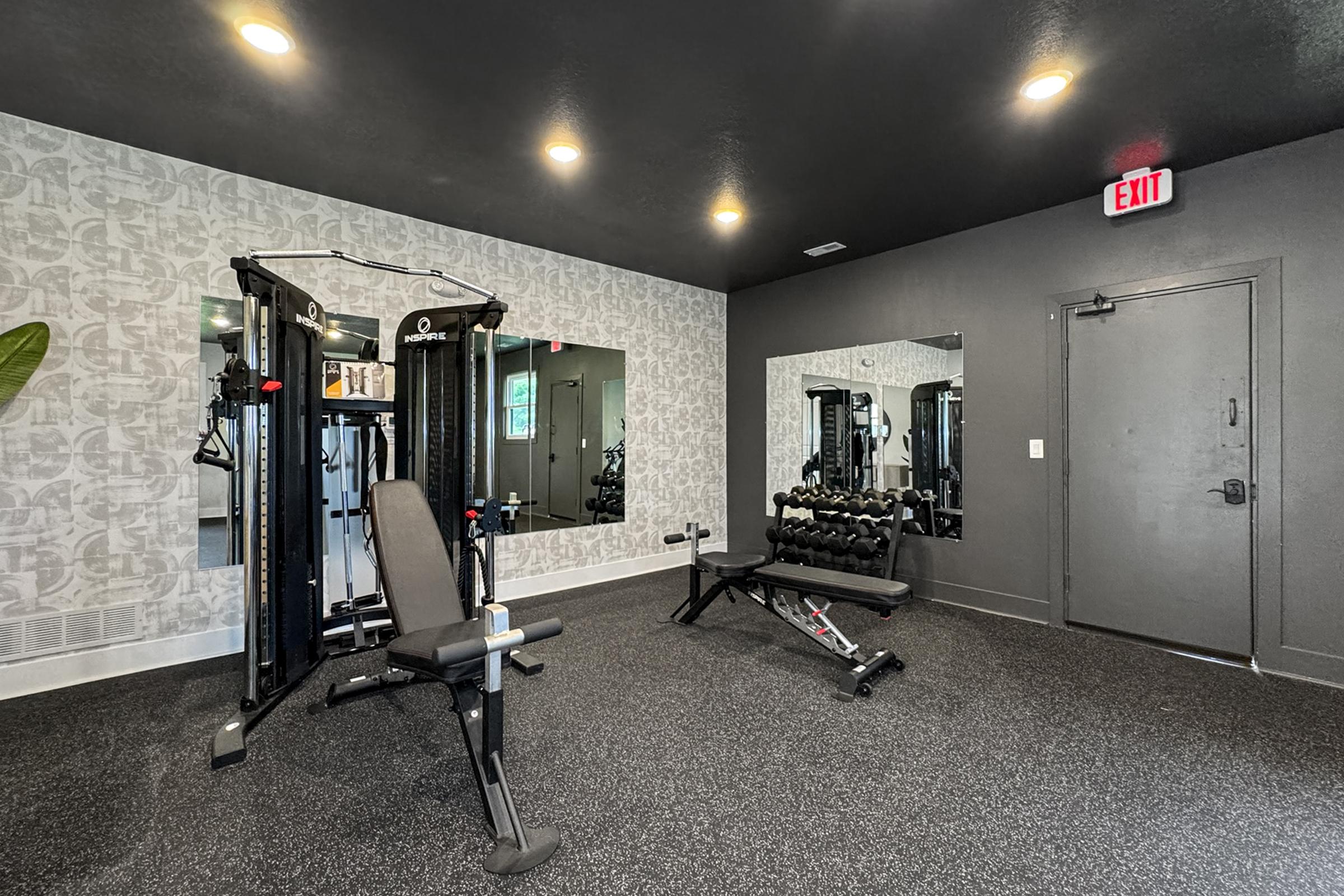 A modern gym interior featuring a multi-function weight machine and a bench press with free weights. The space has dark flooring, a patterned wall, and a large mirror reflecting the equipment. Ambient lighting and an exit sign are visible.