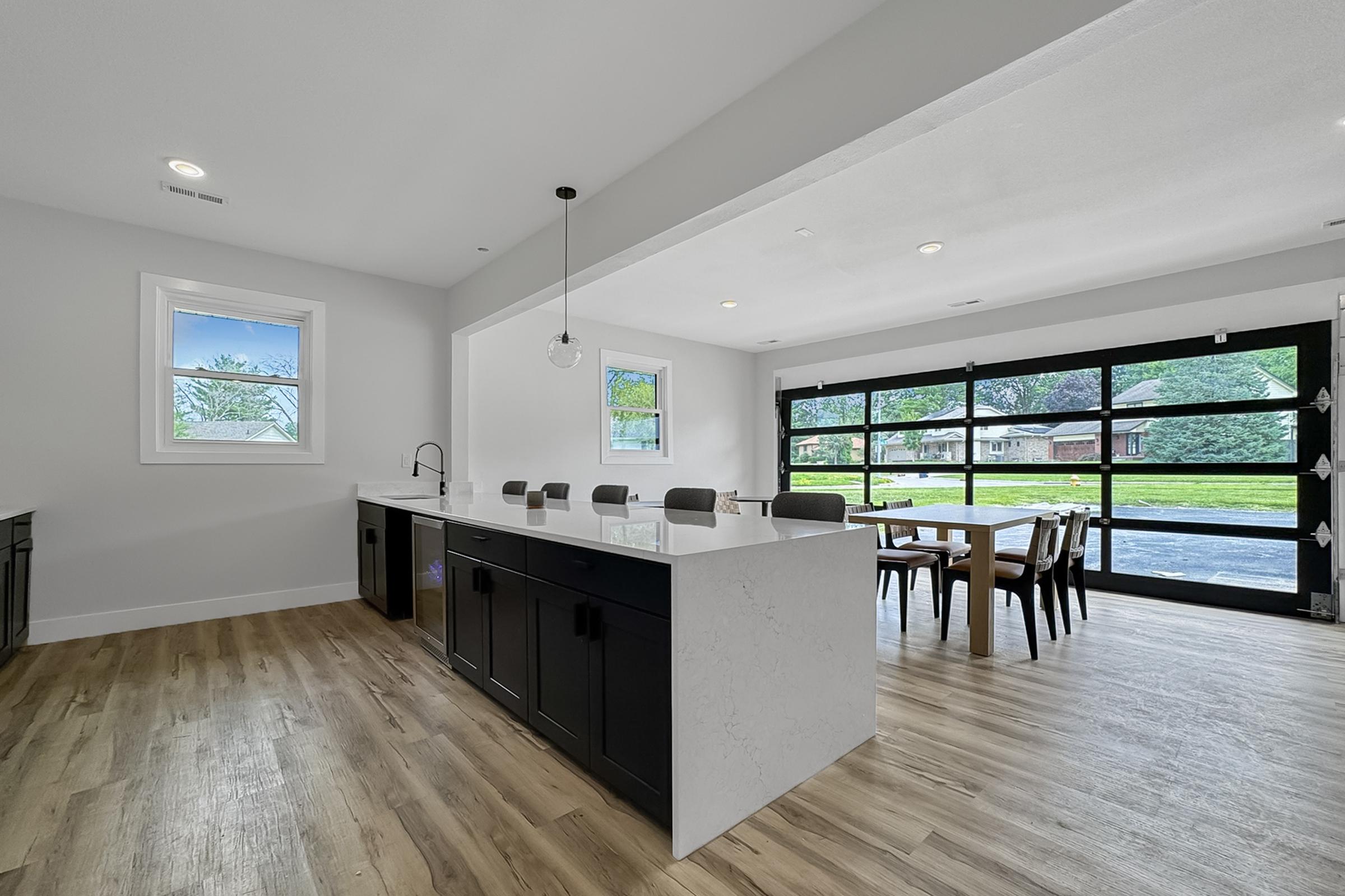 Spacious modern kitchen featuring a large island with seating, sleek black cabinets, and a white countertop. It opens to a dining area with a table set for several people. Large windows provide natural light, and a glass garage door offers a view of the outdoors. Light-colored wood flooring adds warmth to the space.