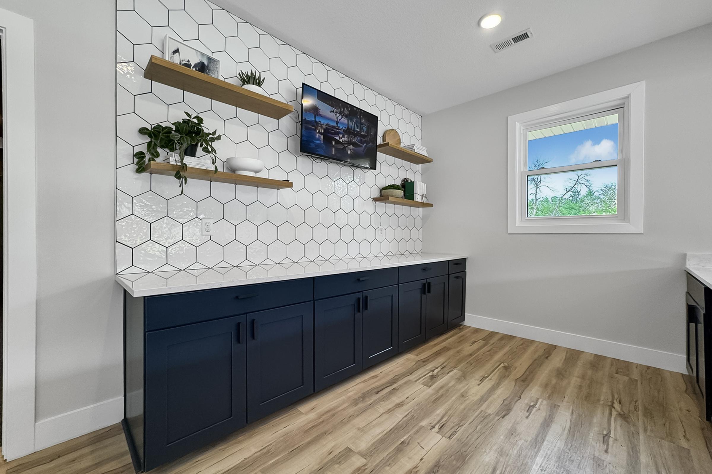 Modern kitchen interior featuring a hexagon-tiled wall with floating wooden shelves displaying plants and decor. A flat-screen TV is mounted on the wall above the shelves. The lower cabinetry is dark blue, and the floor has light wooden planks, complementing the overall contemporary design. A window lets in natural light.