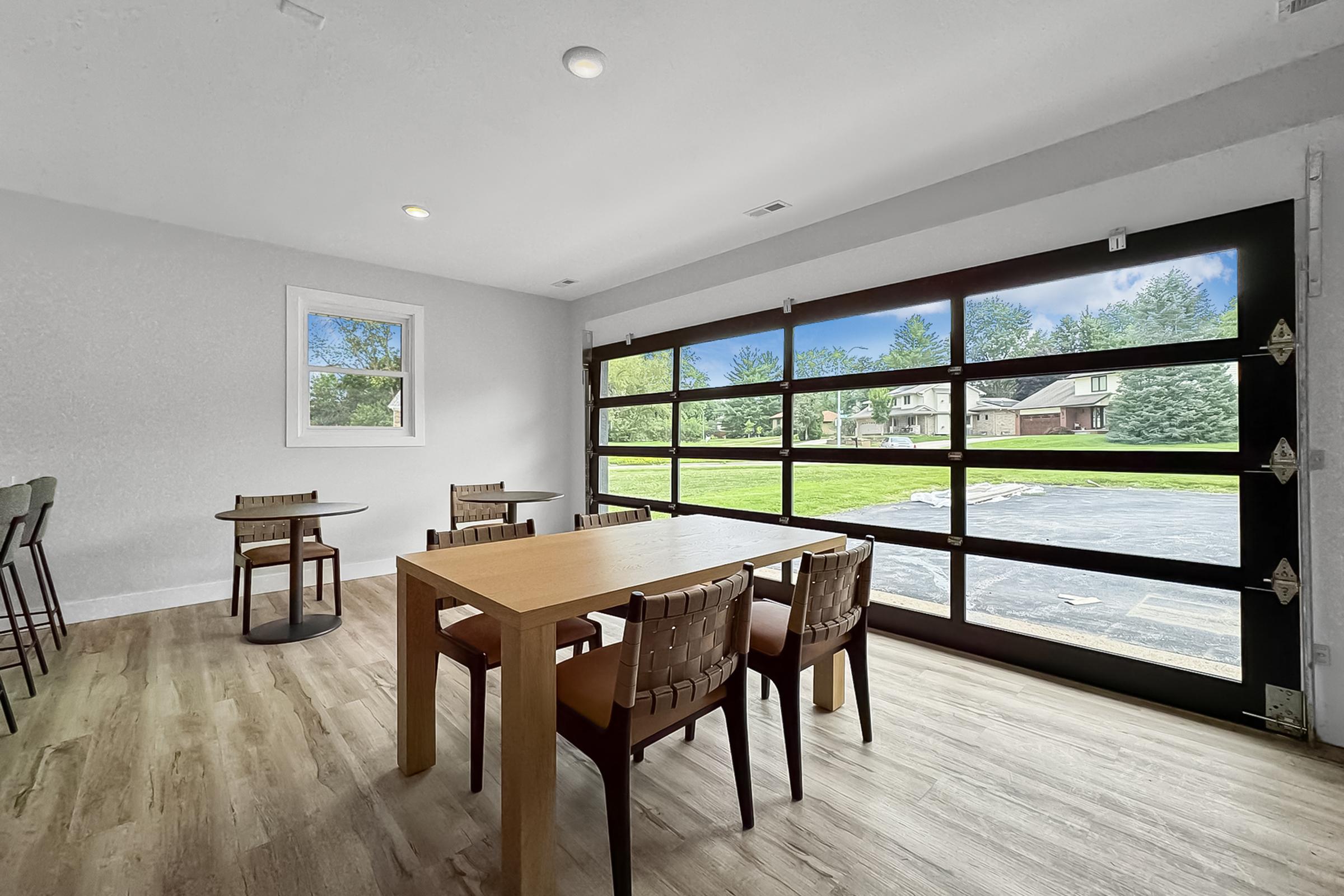 A modern dining area featuring a wooden table surrounded by chairs, with a large glass garage door opening to a view of a green yard and trees outside. The interior has light gray walls and a wooden floor, creating a bright and inviting atmosphere.