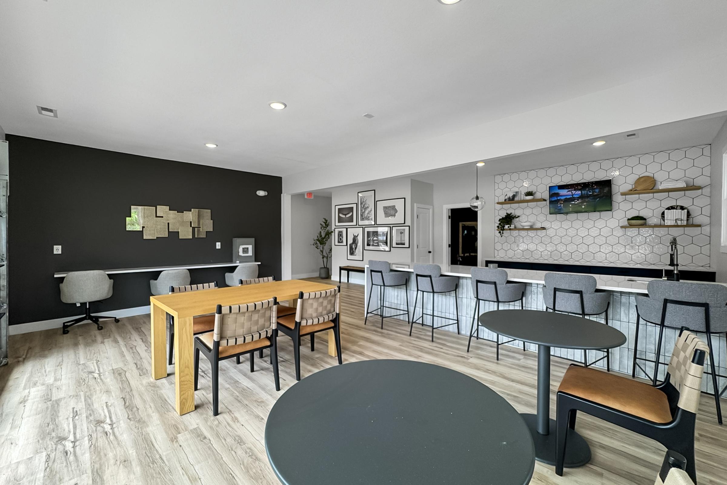 A modern communal space featuring a dining table with wooden benches, round tables with chairs, and a long counter with bar stools. The walls are decorated with framed art, and there's a television mounted above a counter with a geometric design. Natural light illuminates the room.