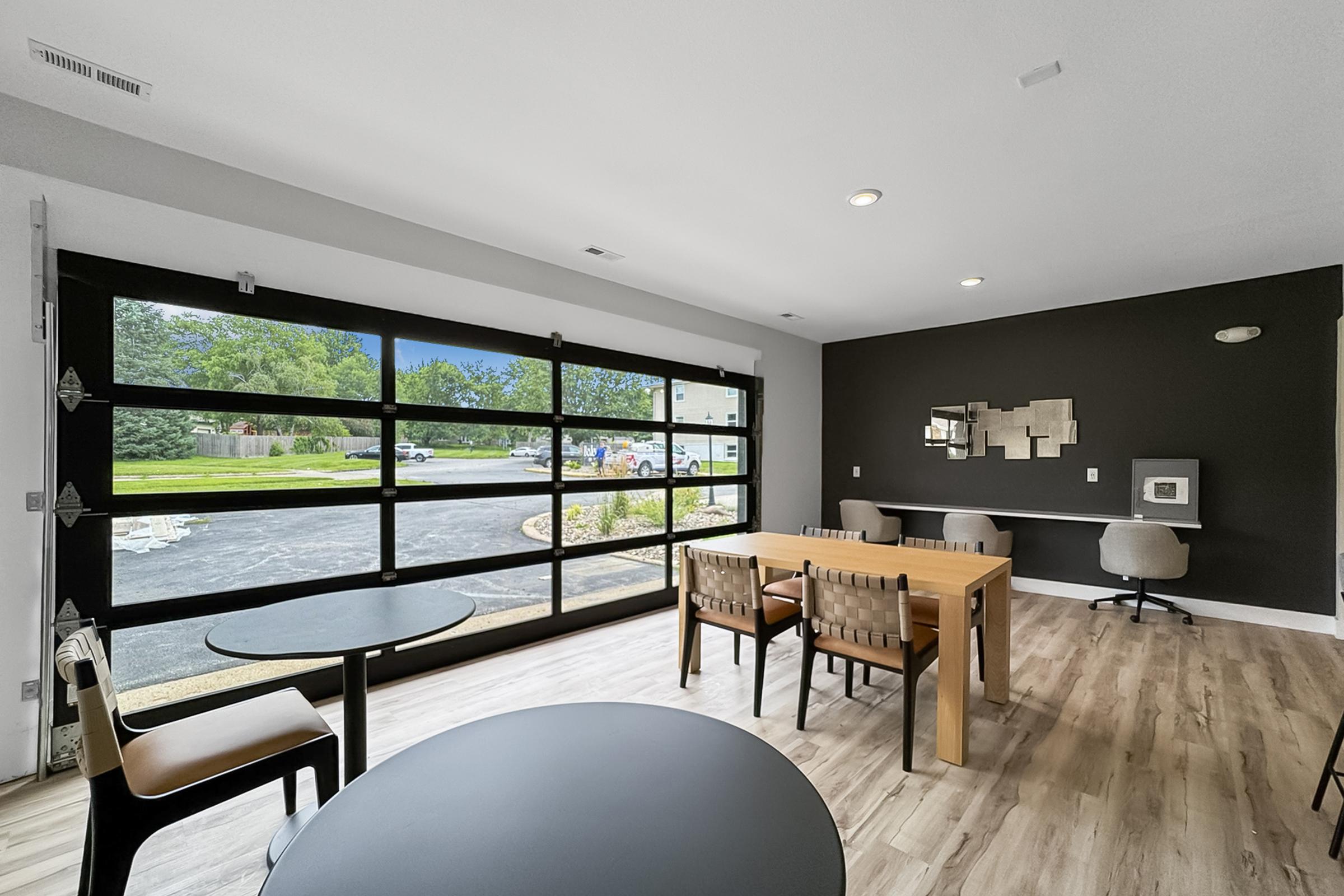 A modern interior space featuring a large glass garage door that opens to an outdoor view. Inside, there are wooden tables and chairs, with a sleek design that includes a dark accent wall and contemporary decor. The area is well-lit with natural light streaming in through the open door.