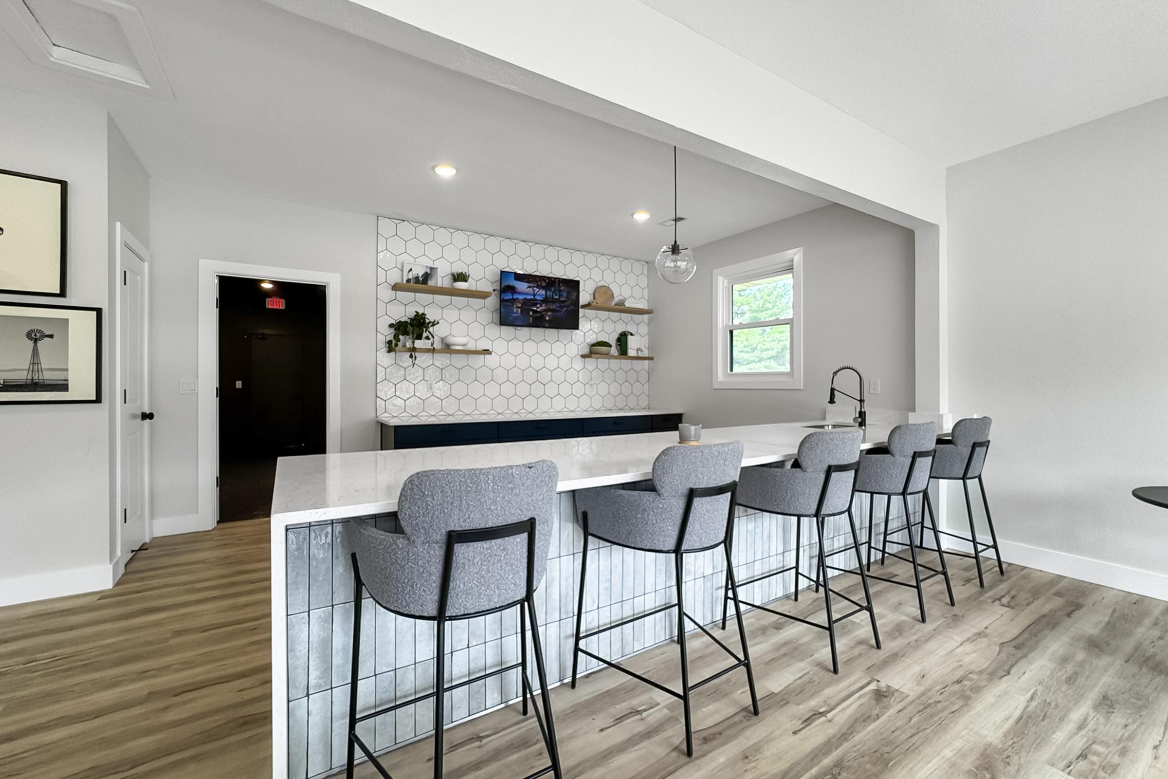 A modern kitchen featuring a large island with gray bar stools, white and gray cabinetry, and a stylish backsplash with hexagonal tiles. In the background, there is a wall-mounted TV and decorative shelves with plants, creating a contemporary and inviting atmosphere.