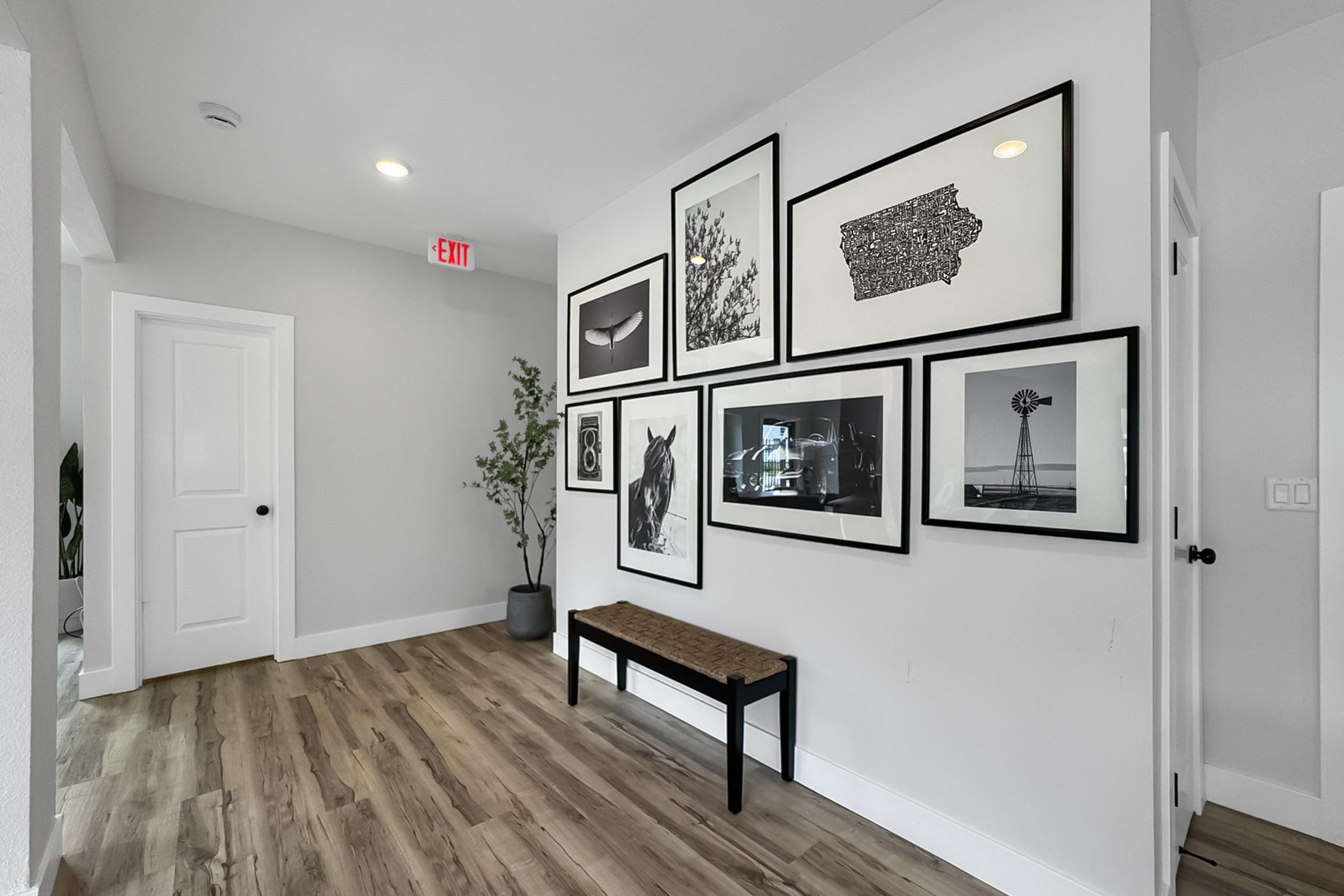 A modern hallway featuring a gallery wall with various framed black-and-white photographs and artwork. A small bench sits on a wooden floor, and a plant adds a touch of greenery. The walls are painted light grey, creating a minimalist and inviting atmosphere. An exit sign is visible above the door.