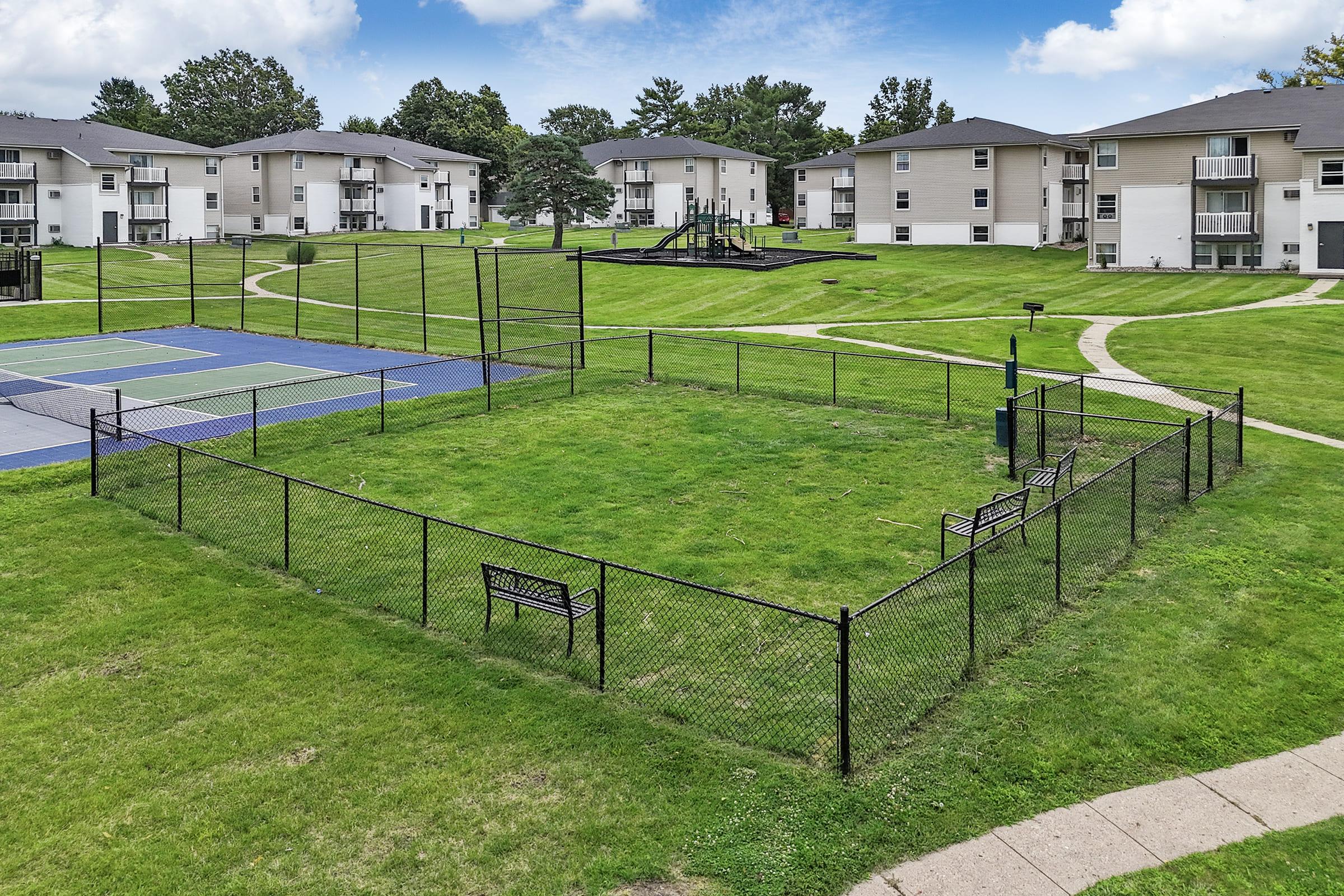 A view of a grassy area enclosed by a black fence, featuring benches and a basketball hoop. Nearby, there are tennis courts and a playground, with residential buildings in the background. The scene is set in a well-maintained outdoor space with blue skies and scattered clouds.