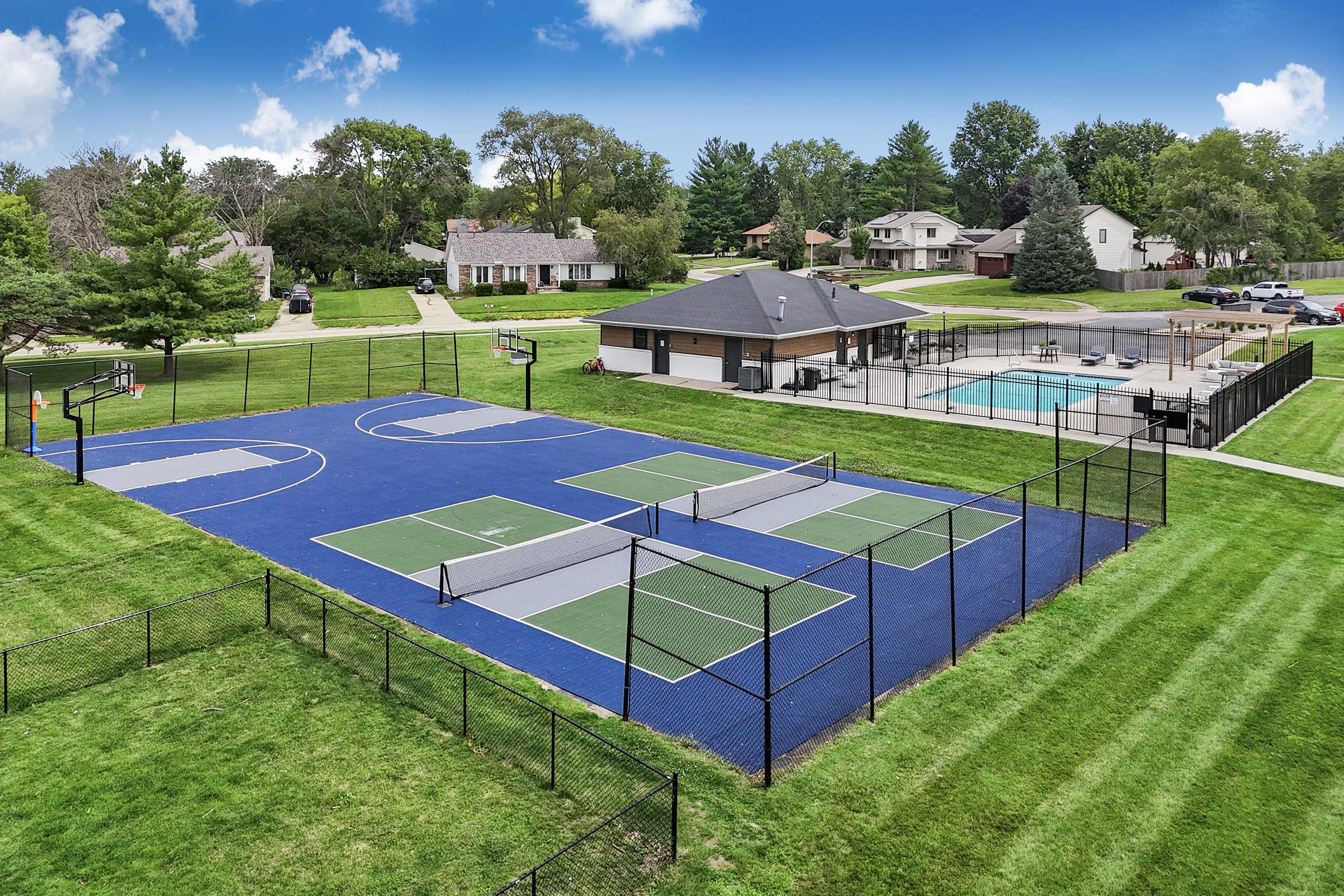 Aerial view of a sports complex featuring a multi-purpose court with basketball and tennis sections, surrounded by green lawns. A swimming pool is visible nearby, along with residential homes in the background. Clear blue sky adds to the inviting atmosphere of the recreational area.