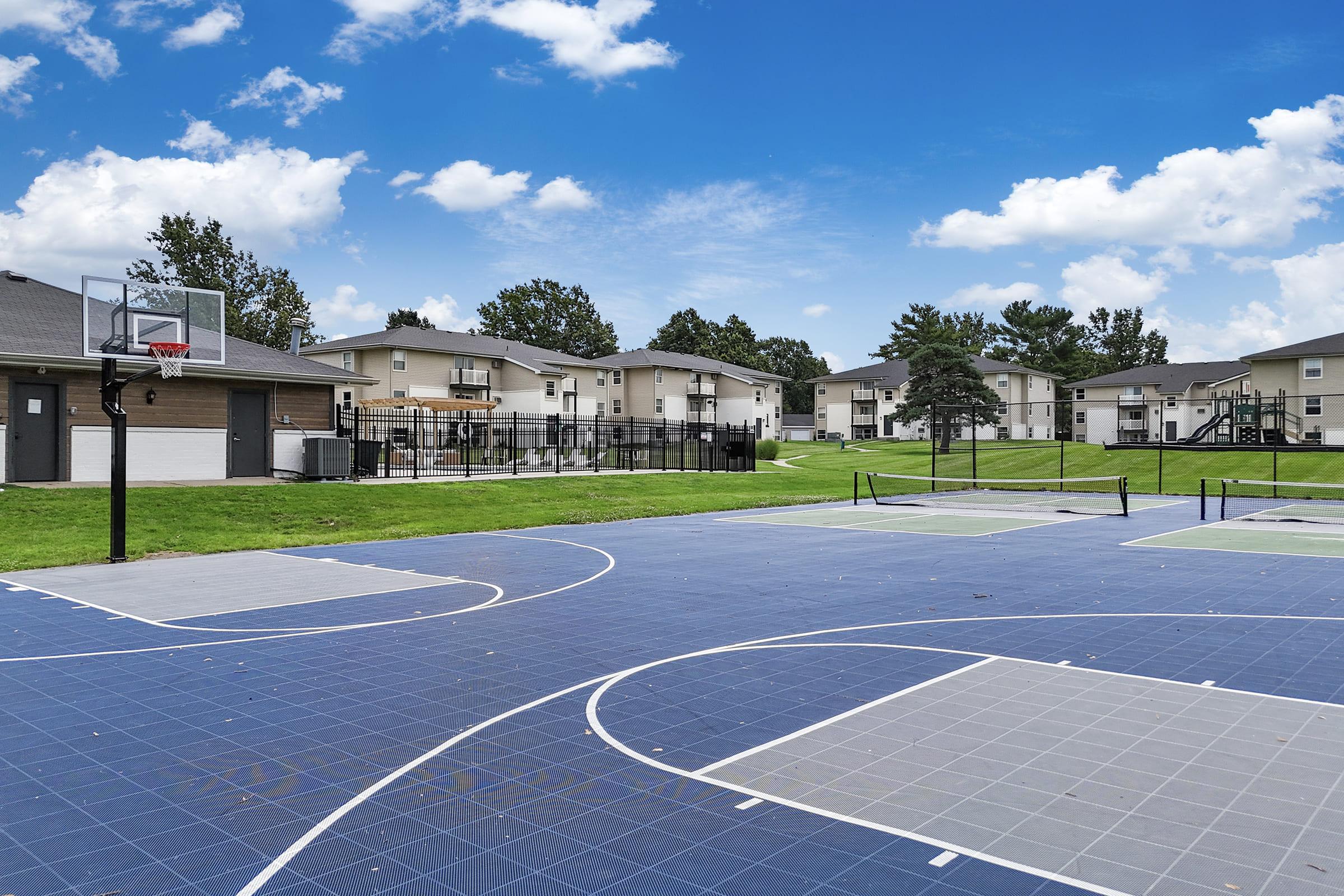 View of a basketball court with a blue surface, surrounded by green grass and several apartment buildings in the background. The court features basketball hoops and is set against a bright blue sky with scattered clouds.