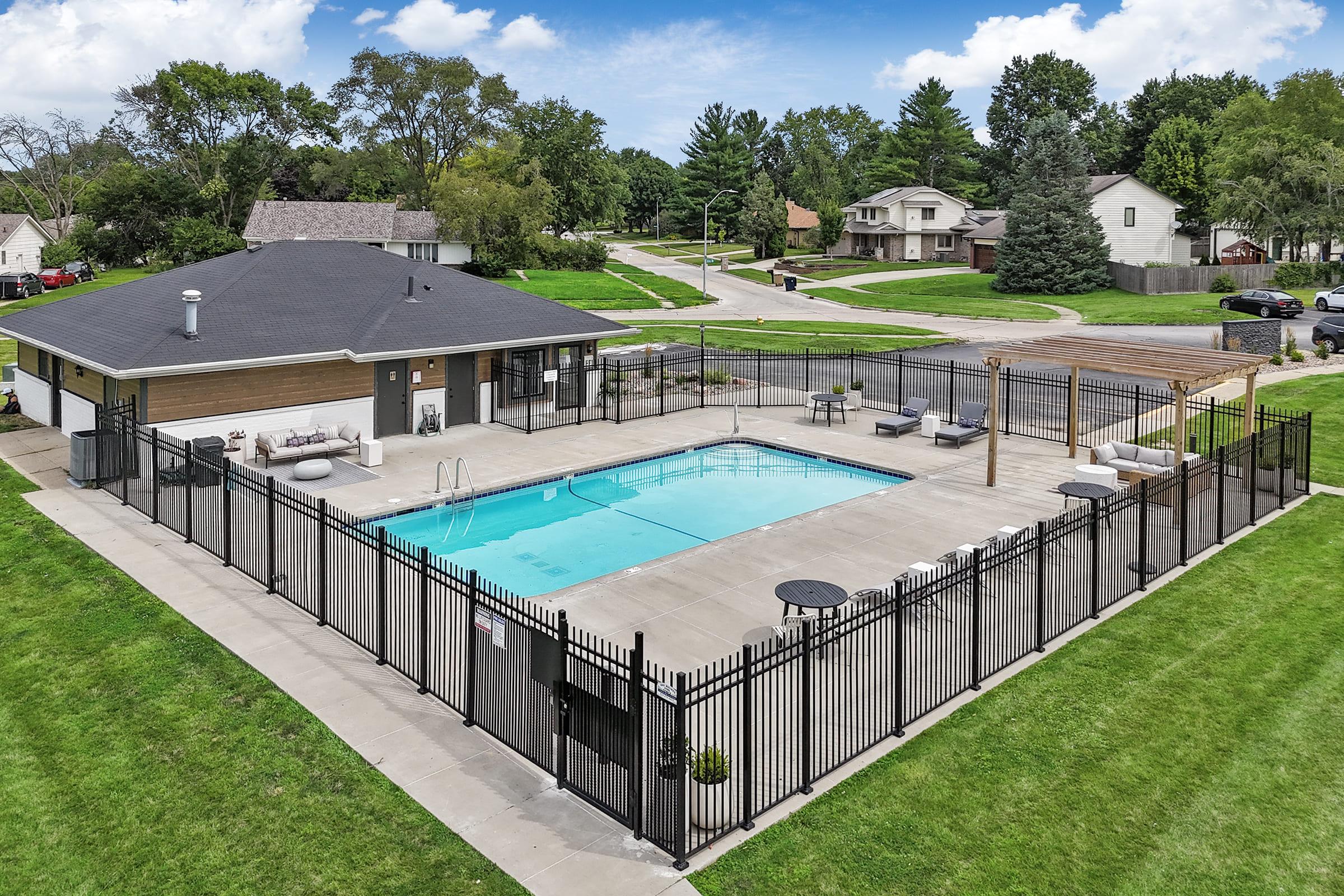 Aerial view of a fenced outdoor swimming pool surrounded by green grass and trees. The pool area features lounge chairs, tables, and a covered seating area, with a residential neighborhood visible in the background. Clear blue sky and well-maintained landscaping add to the inviting atmosphere.