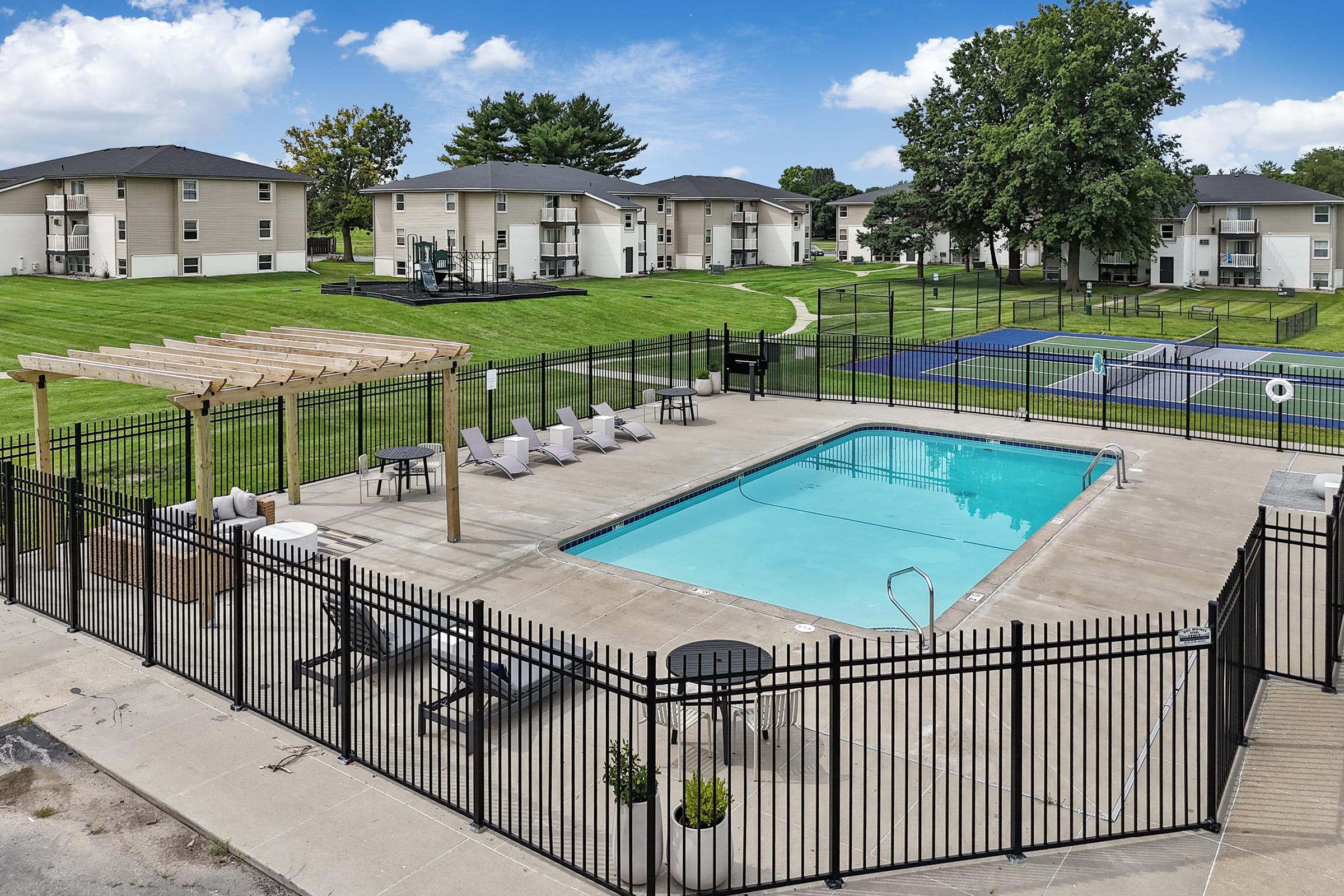 A serene outdoor pool area featuring a clear blue pool surrounded by lounge chairs, a pergola for shade, and a fenced perimeter. In the background, there are residential buildings and a tennis court, set against a backdrop of green grass and trees under a partly cloudy sky.