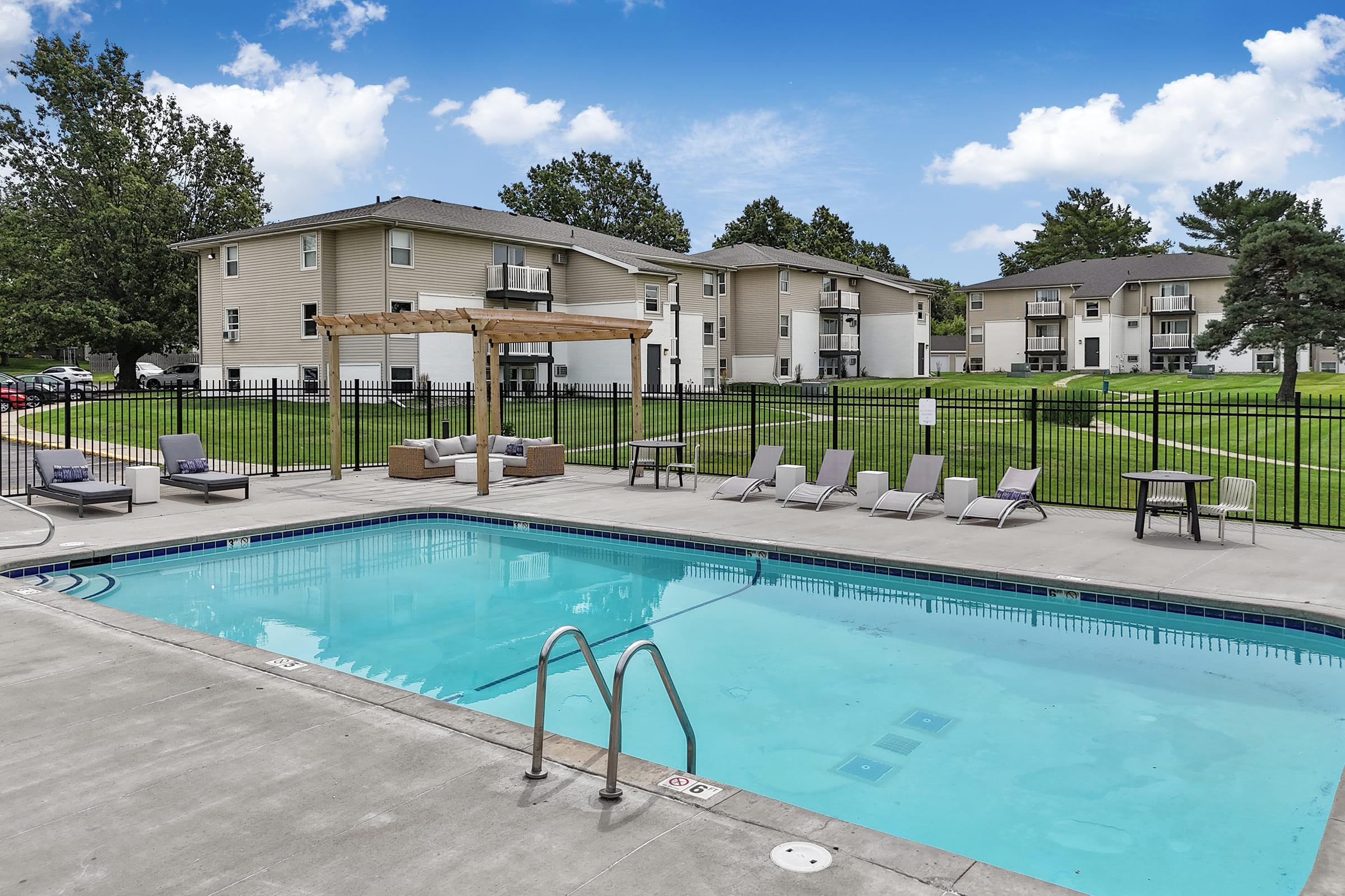 A tranquil outdoor swimming pool with lounge chairs and a shaded pergola. Surrounding green lawns and residential buildings in the background, set against a clear blue sky, create a serene atmosphere for relaxation and leisure.