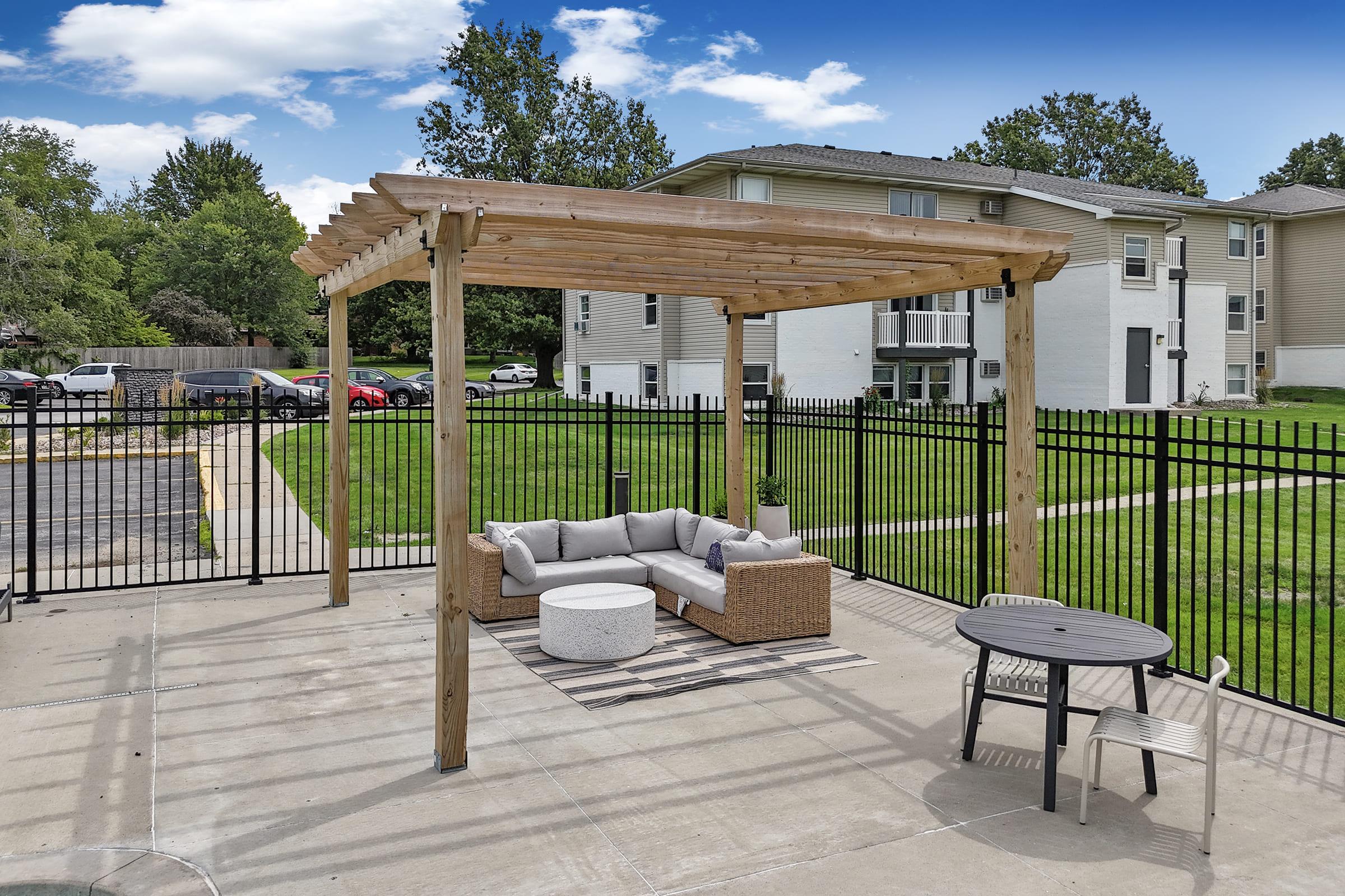 A cozy outdoor seating area featuring a wooden pergola with a comfortable couch and a round coffee table, set on a paved patio. In the background, there's a well-maintained grassy area and apartment buildings, with a fenced perimeter and a parking lot. Bright blue sky and scattered clouds add to the scenic view.