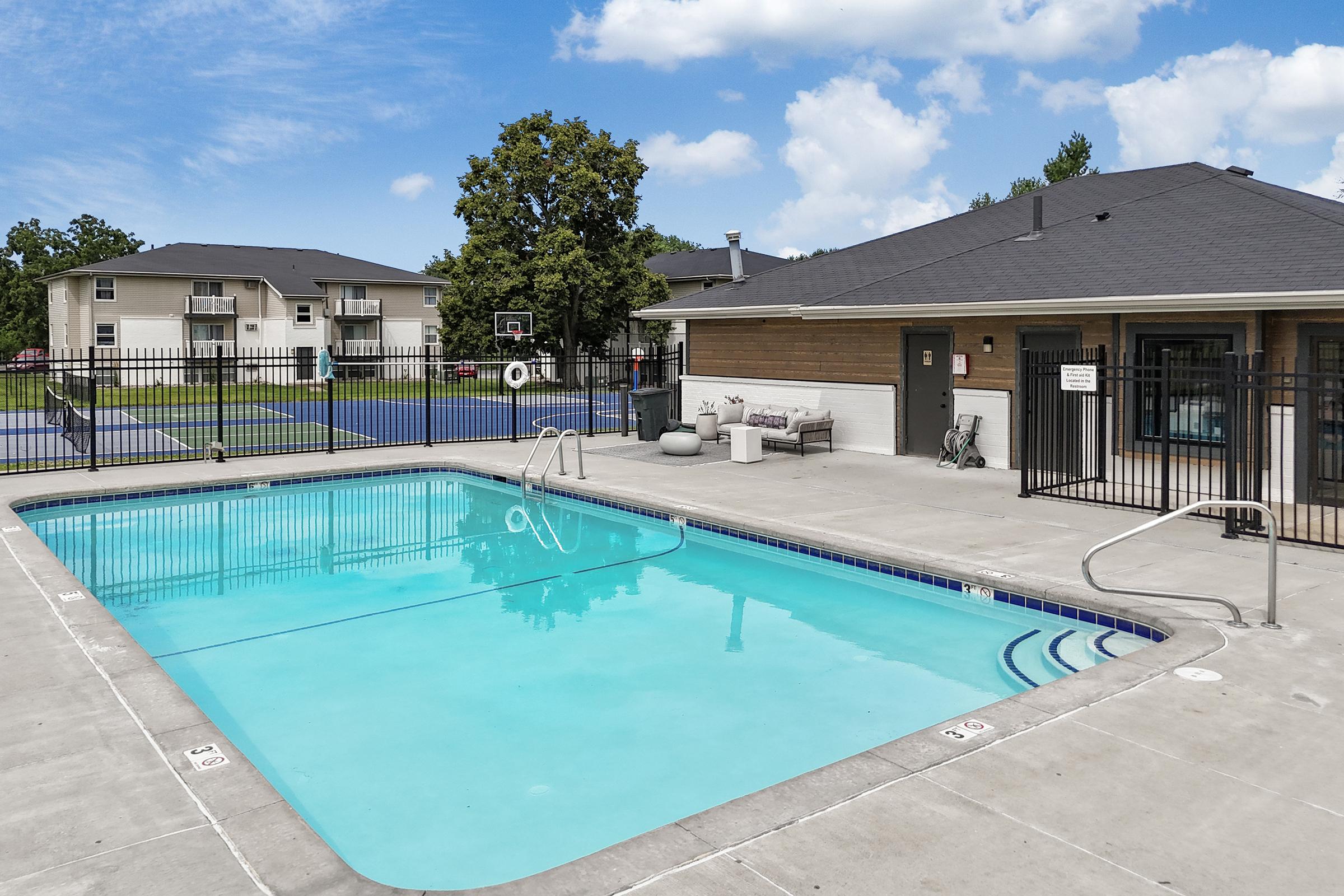 A clear blue swimming pool surrounded by a concrete area, with steps leading into the water. Nearby, there are lounge chairs and a fenced patio. In the background, there are residential buildings and a basketball court. The sky is bright with scattered clouds.