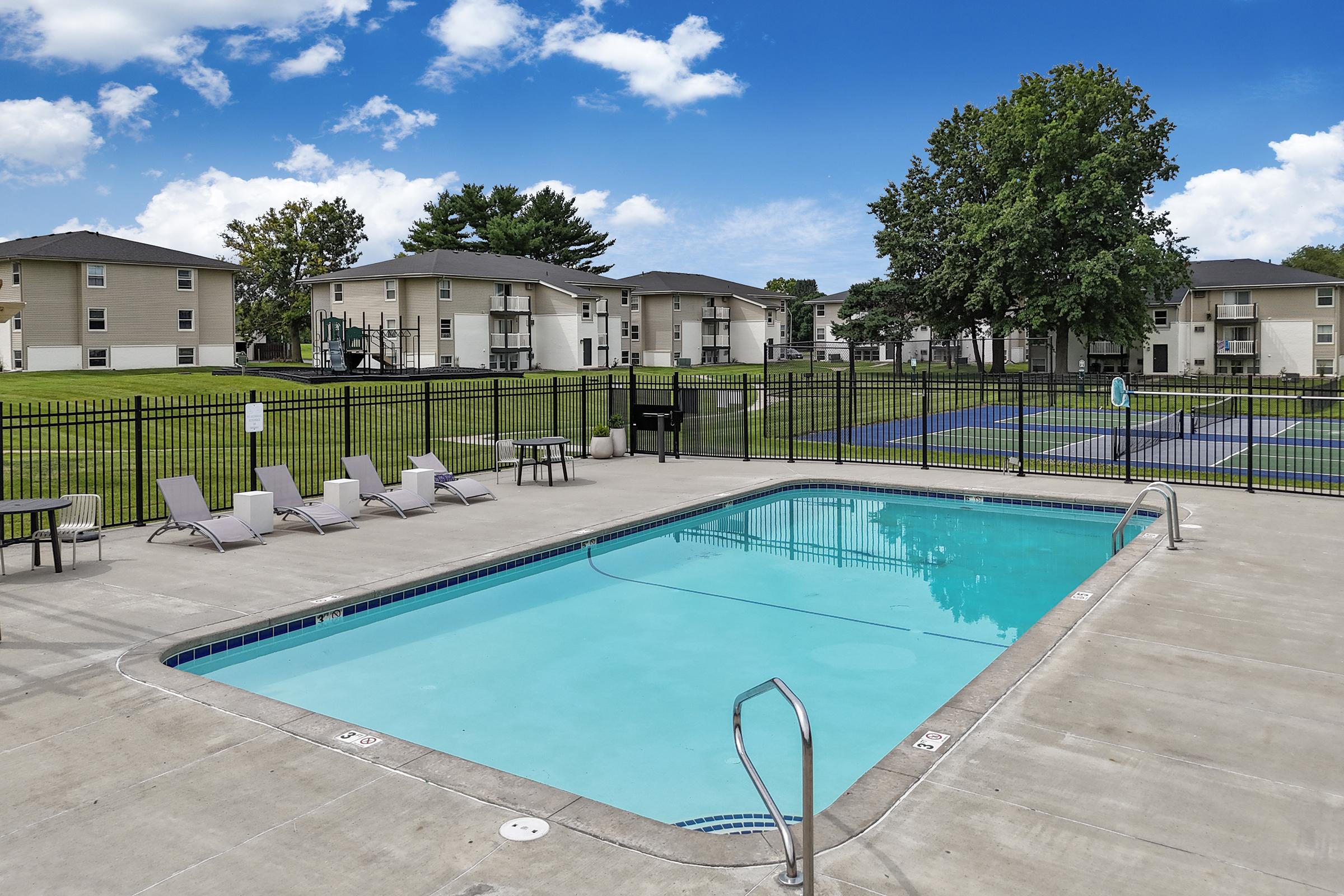 A swimming pool with clear blue water surrounded by gray pavement. Lounge chairs are positioned by the poolside. In the background, there are multi-story residential buildings and a tennis court. The sky is bright with white clouds, creating a vibrant atmosphere.