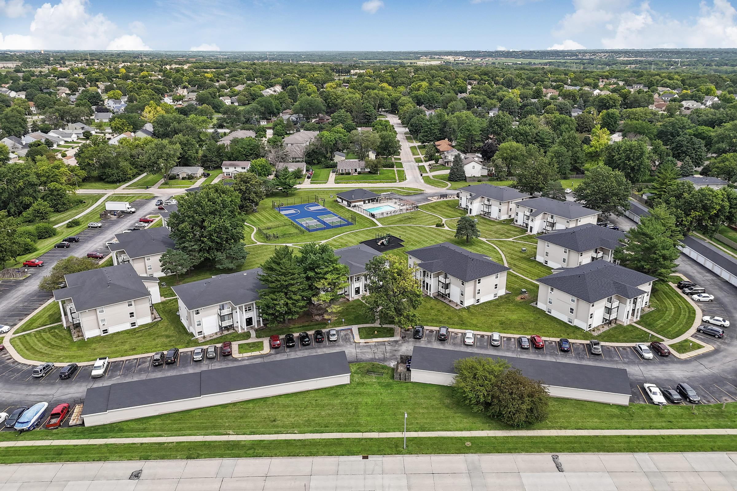Aerial view of a residential apartment complex surrounded by green lawns and trees. The complex features multiple buildings arranged in a circular layout, a swimming pool, and recreational areas. Roads and parking spaces are visible, along with houses and greenery in the surrounding area.
