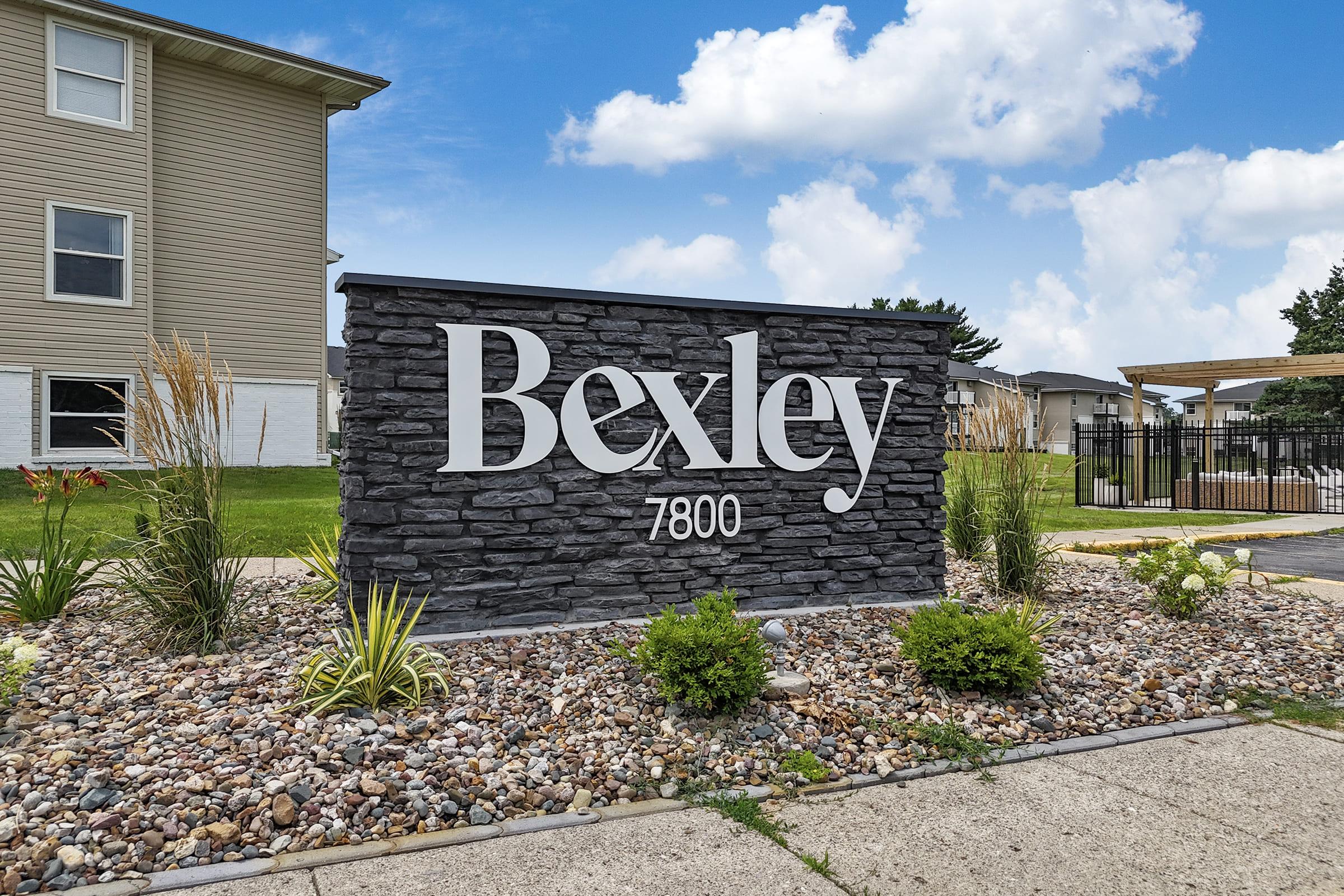 A stone sign displaying the word "Bexley" with the number "7800" below it. The sign is surrounded by decorative landscaping, including small plants and rocks, set against a backdrop of a grassy area and blue sky with clouds.