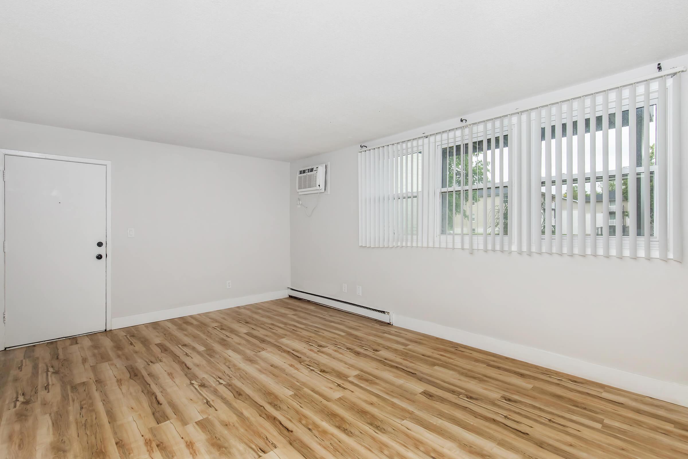 Bright living space with air conditioning unit, vertical blinds, and updated flooring in Bexley 1-bedroom apartment.