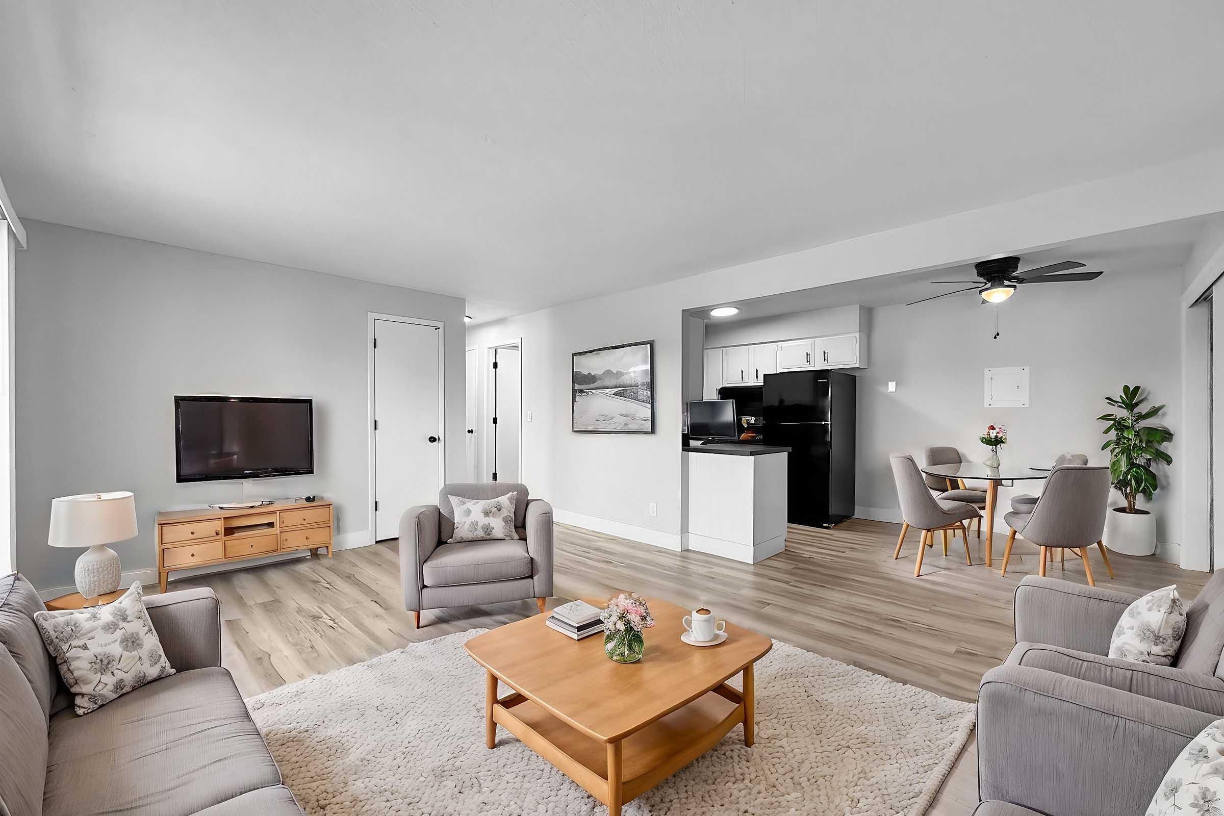 A bright and modern living room featuring a light grey sofa and armchairs, a wooden coffee table with a flower vase, and a television on a wooden unit. The open kitchen is visible in the background, with black appliances and a dining area with a round table and chairs. Light-colored walls and flooring create a spacious atmosphere.