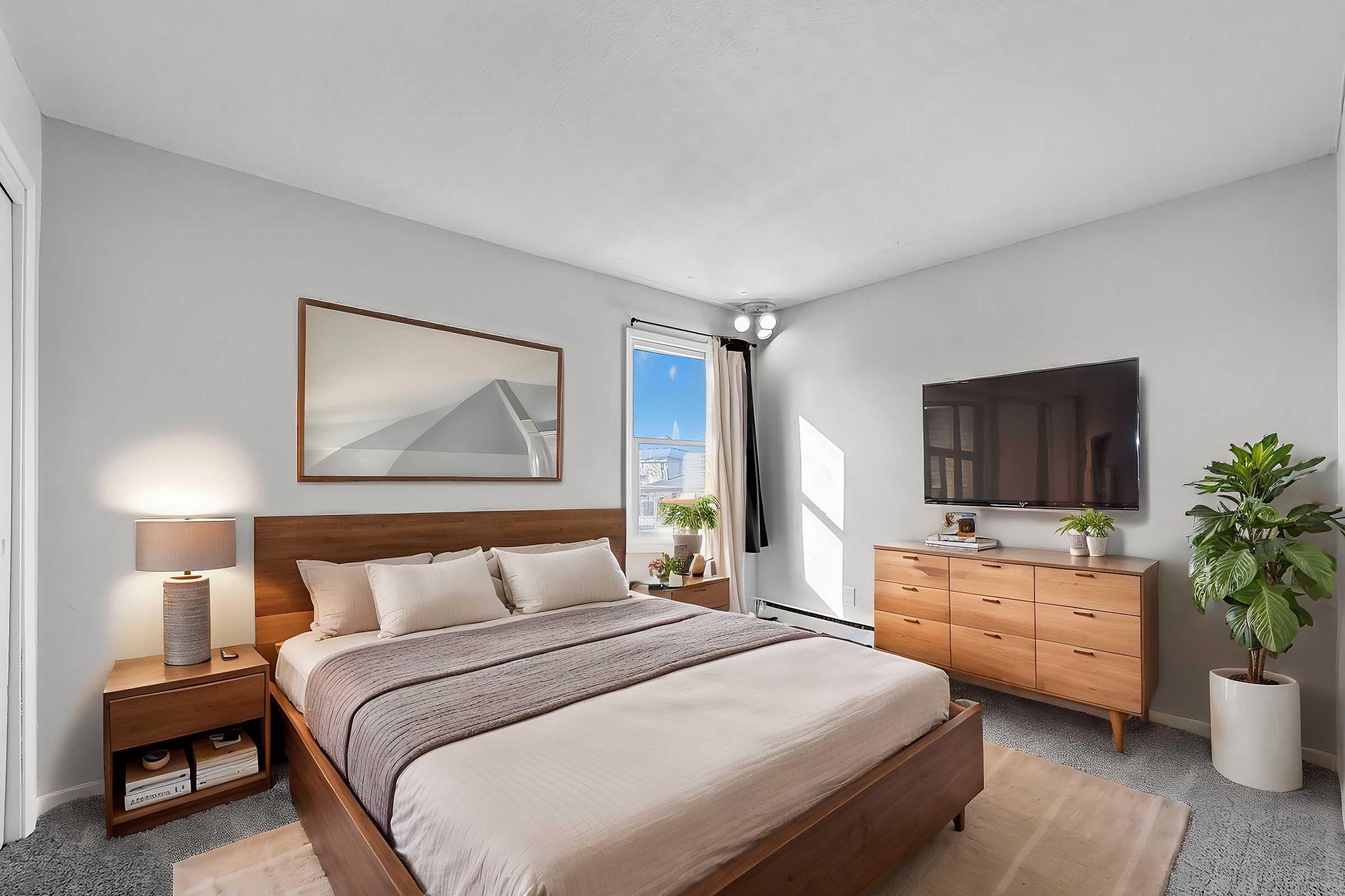 A modern bedroom featuring a wooden bed with neutral bedding, a stylish lamp on a nightstand, a large flat-screen TV mounted on the wall, and a wooden dresser. Natural light streams in through a window, while a potted plant adds a touch of greenery to the space. The overall decor is minimalist and inviting.
