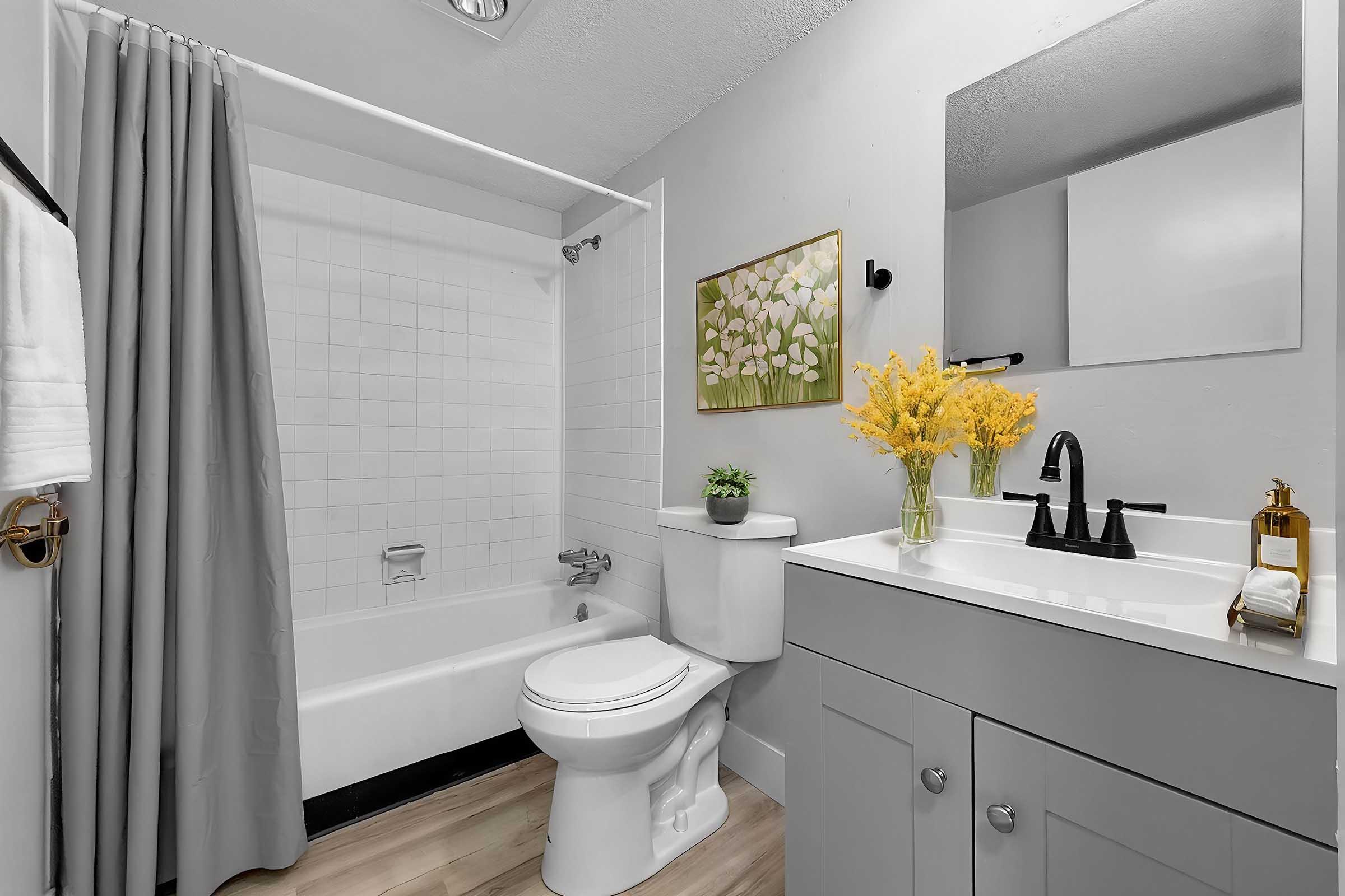 A modern bathroom featuring a white bathtub with a gray curtain, a white toilet, and a gray vanity with a sink. A small plant and a vase with yellow flowers adorn the vanity. The walls are painted light colors, and there is a floral painting on the wall. The floor has light wood tiles.