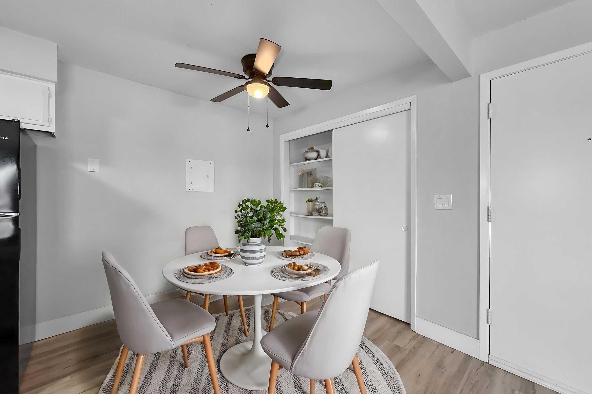 A modern dining area featuring a round white table set with plates of food, surrounded by four light gray chairs. A ceiling fan is visible above, and there is a small plant on the table. The walls are painted in a light gray color, and a built-in cabinet is partially visible in the background.