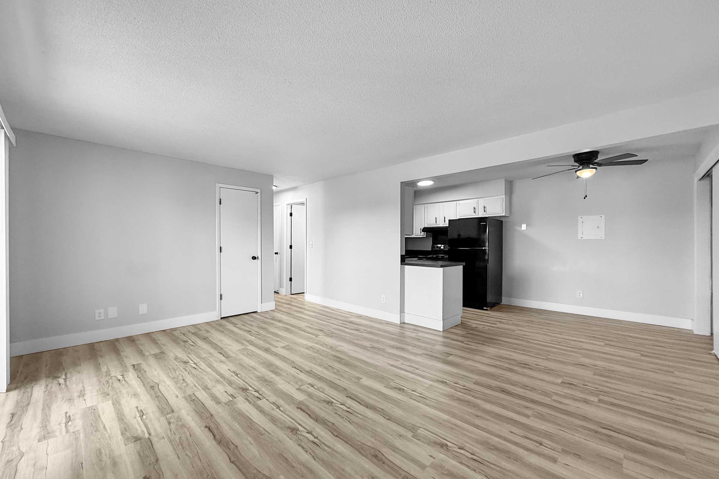 A spacious, modern living area featuring light grey walls, a ceiling fan, and laminate flooring. A kitchen area with black appliances is visible in the background, and a doorway leads to another room. Natural light brightens the open layout, giving a fresh and inviting atmosphere.