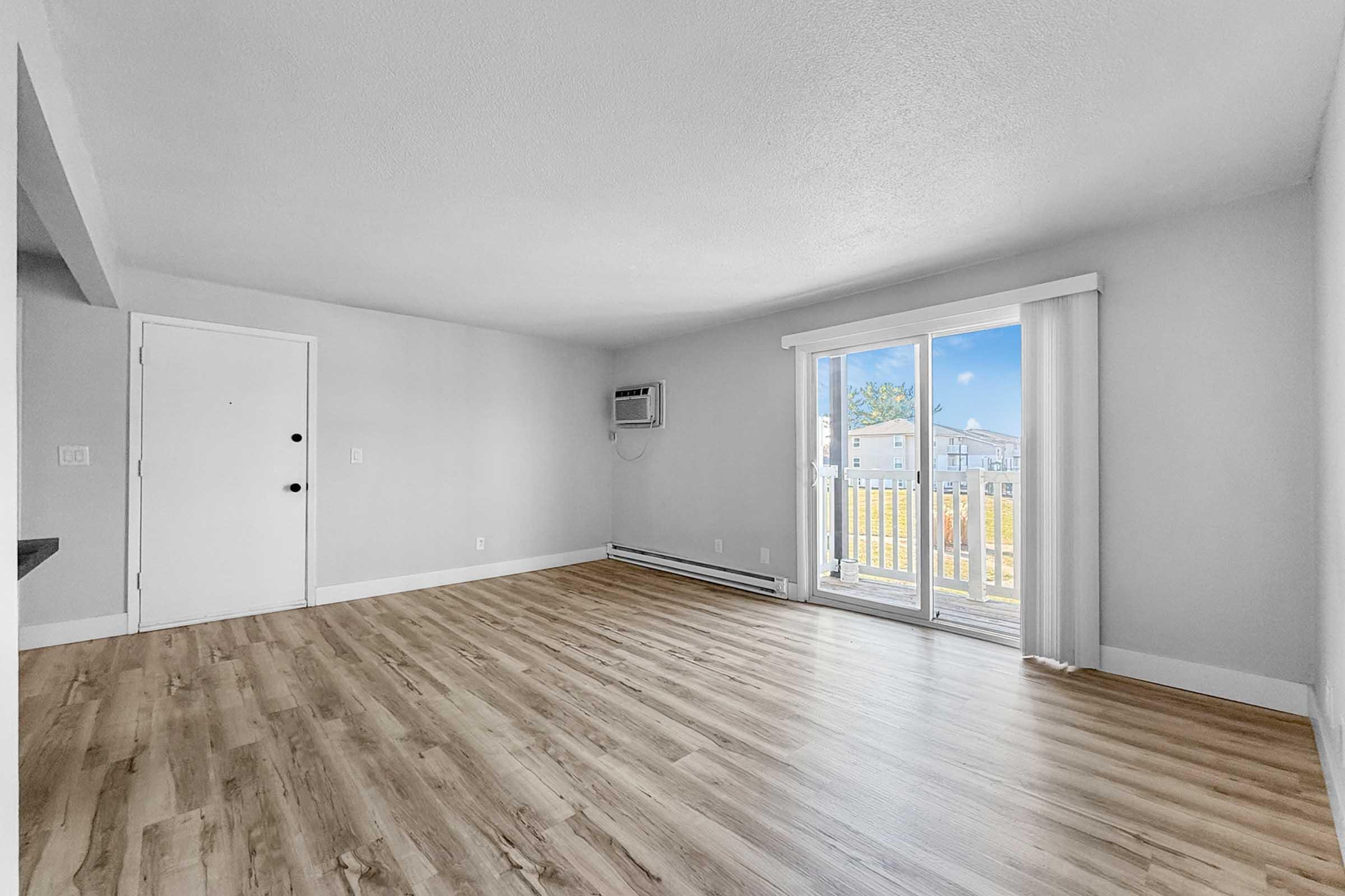 Spacious, well-lit living room featuring light wood flooring, white walls, and a sliding glass door leading to a small balcony. An air conditioning unit is visible on the wall, and a door is visible on the left side. Natural light comes in from the outside.