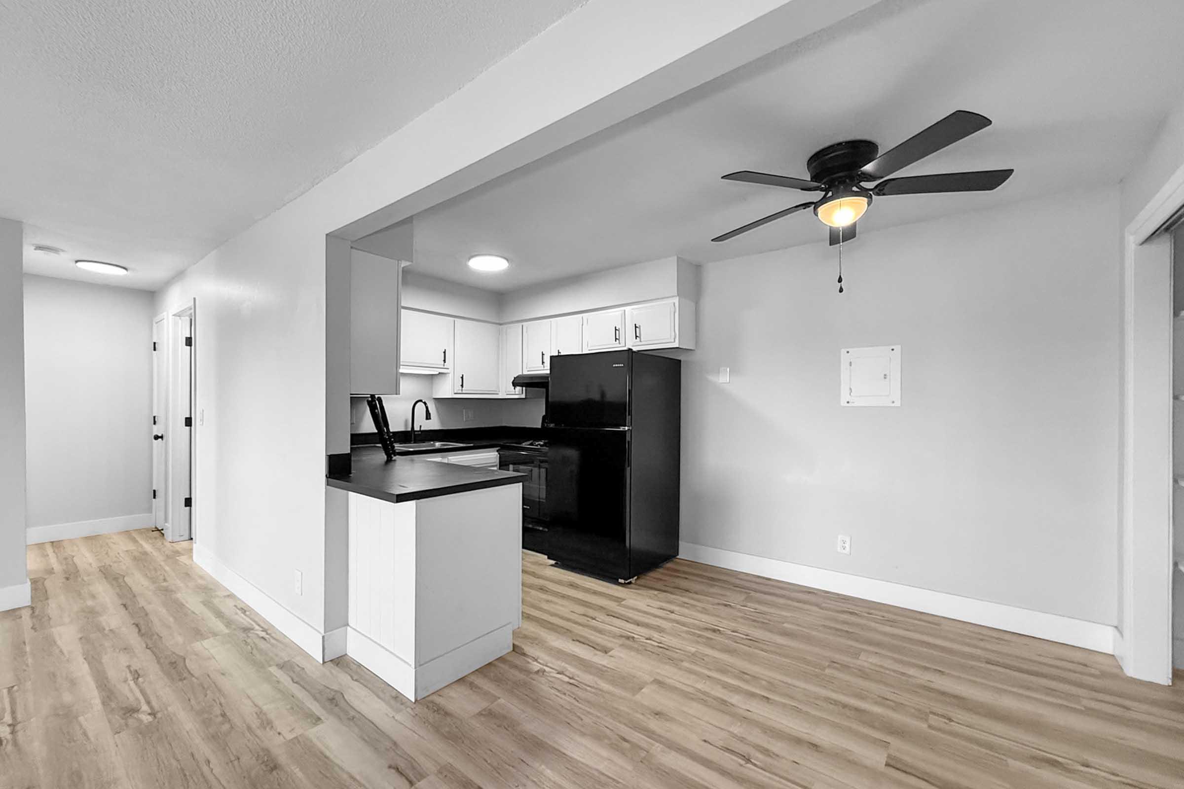A modern kitchen featuring a black refrigerator and sleek black countertops, with white cabinetry. A ceiling fan is visible, and the flooring is light-colored wood. The space is well-lit with recessed lighting and has an open layout leading to an adjacent room.