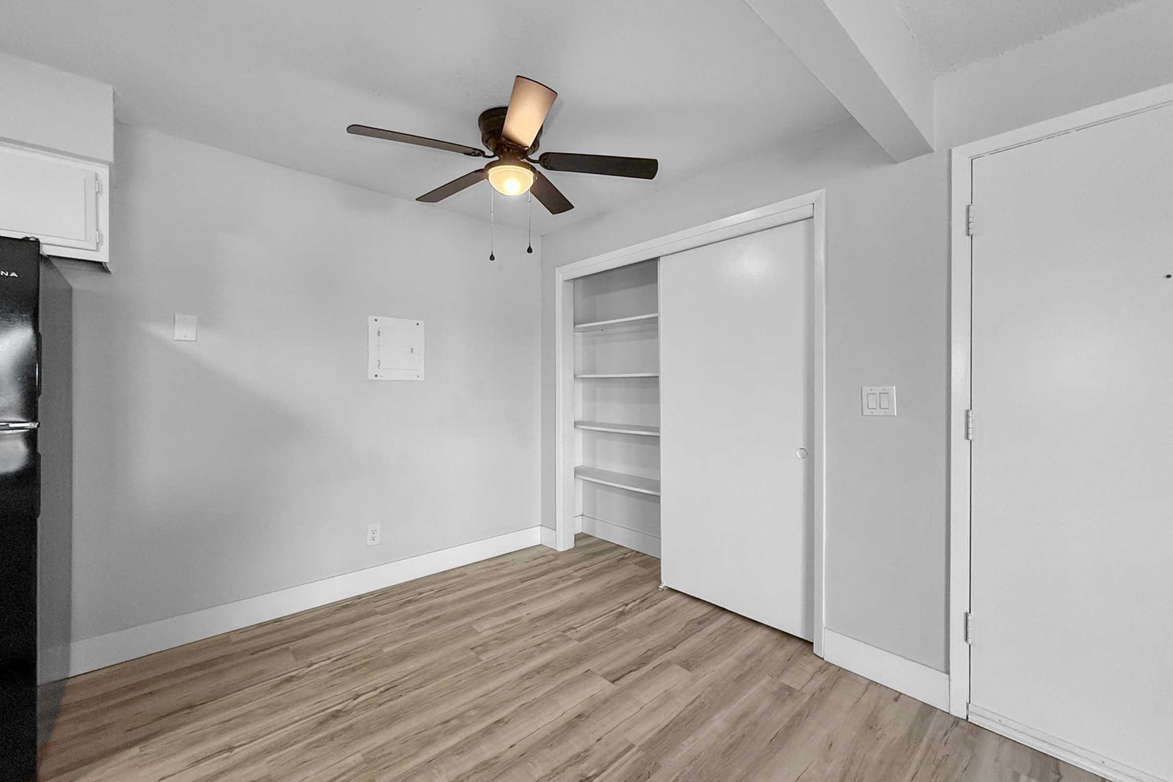 A minimalist room featuring a ceiling fan, light wood-style flooring, and a white closet with sliding doors. The walls are painted in a light gray color, and a black refrigerator is visible in the corner. The design emphasizes simplicity and functionality, with a clean and modern aesthetic.