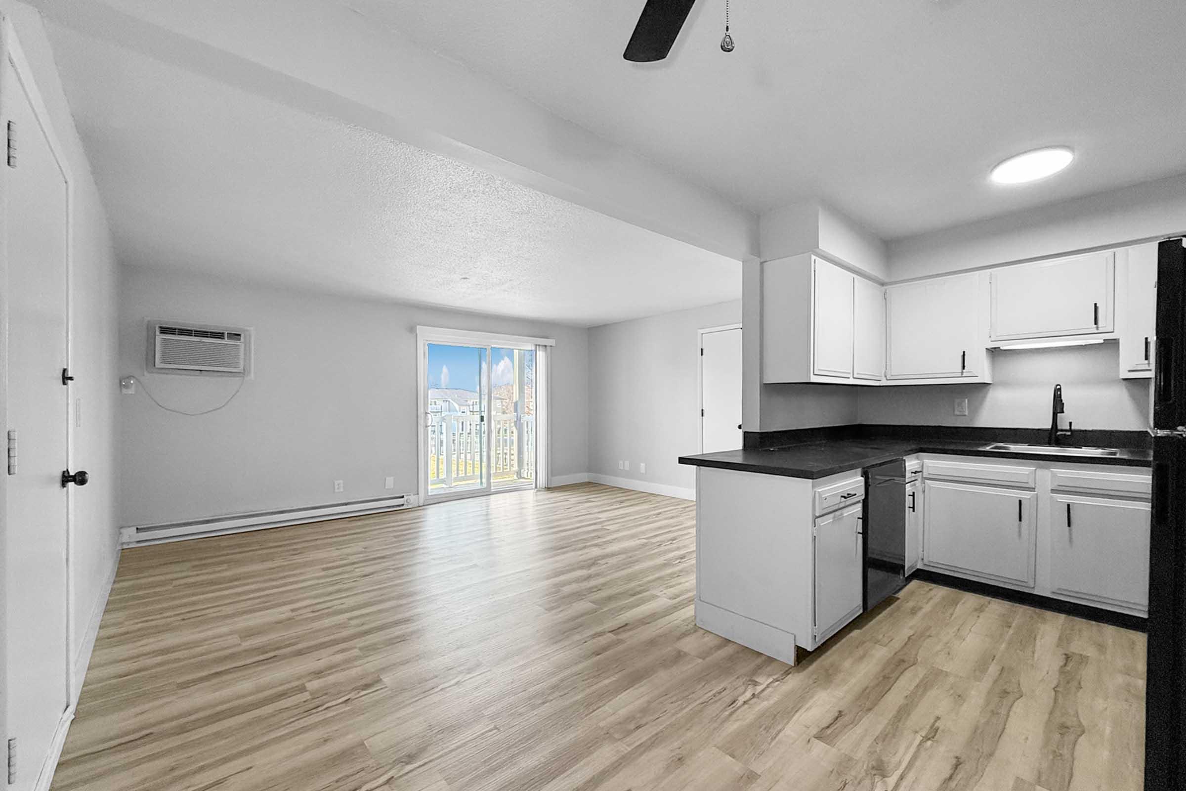 A bright and spacious room featuring an open kitchen area with white cabinets, a black countertop, and stainless steel appliances. The living space has light-colored wooden flooring and large windows allowing natural light; a sliding glass door leads to an outdoor balcony.